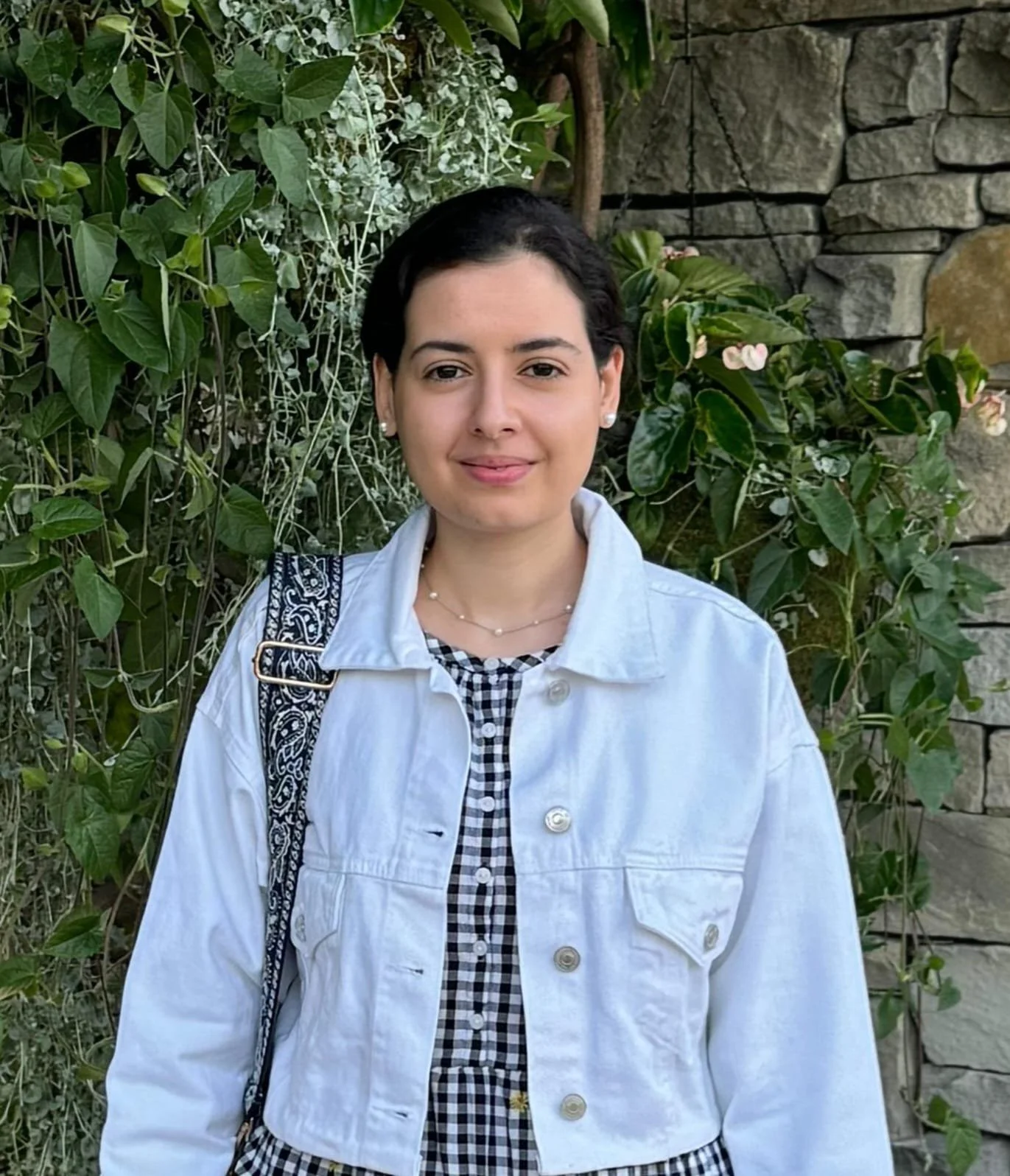 A woman with dark hair, wearing a white denim jacket over a black and white checkered dress, standing outdoors near a stone wall surrounded by green plants and flowers.