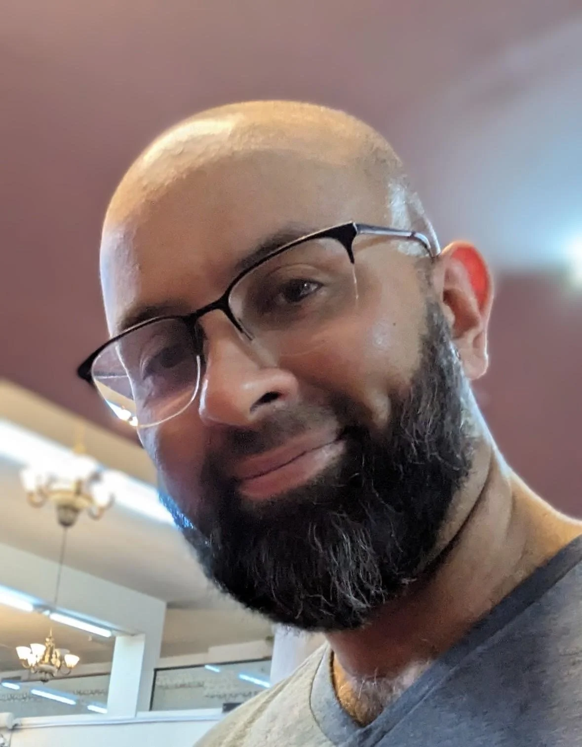 A close-up of a man with a bald head, glasses, a beard, and a mustache, smiling in an indoor setting.