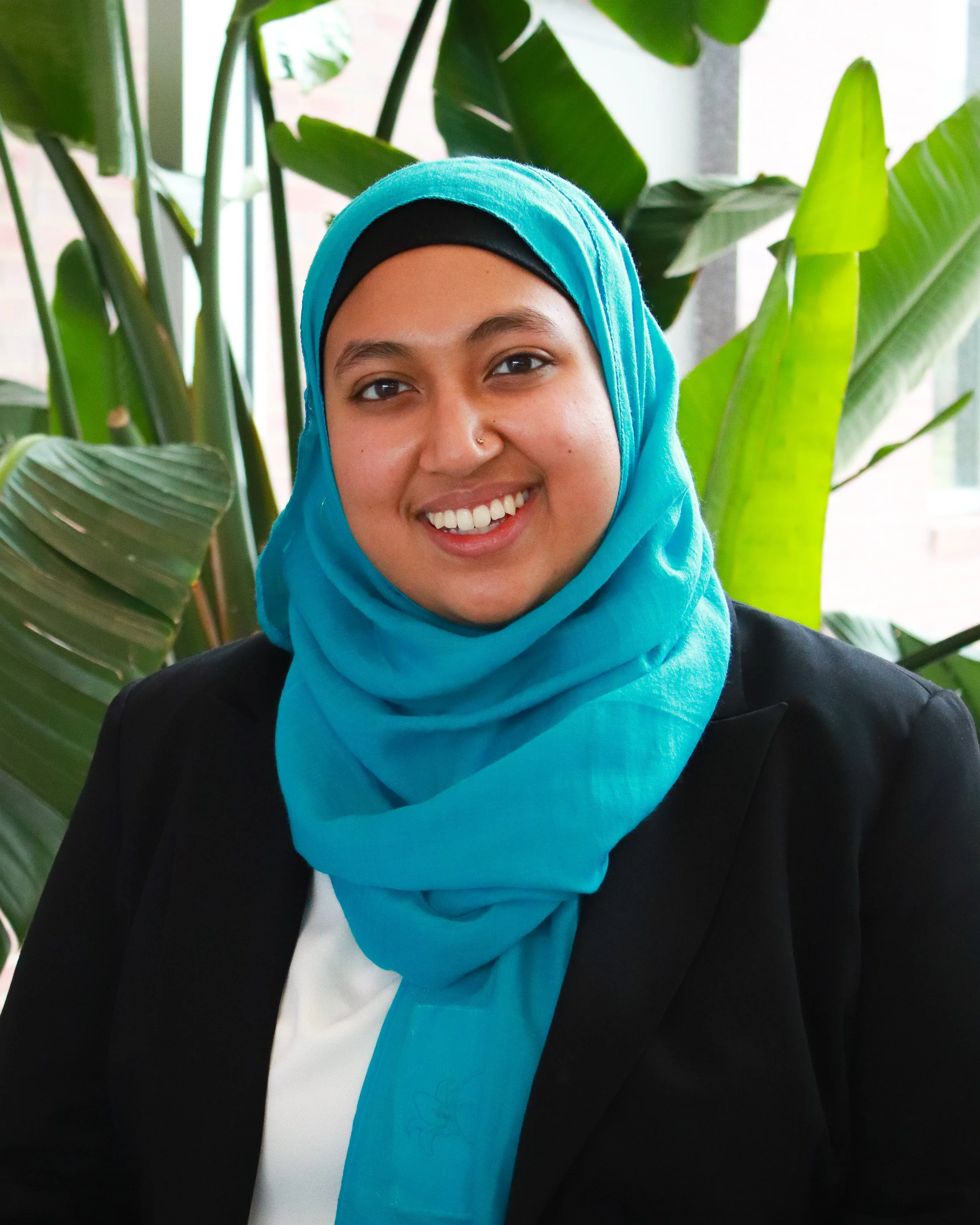 A woman smiling, wearing a blue headscarf and a black blazer, with large green plants in the background.