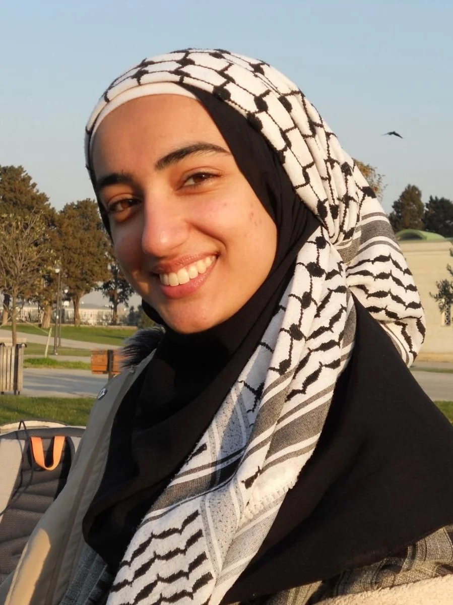 A woman wearing a black and white patterned headscarf smiling outdoors in a park during sunset.