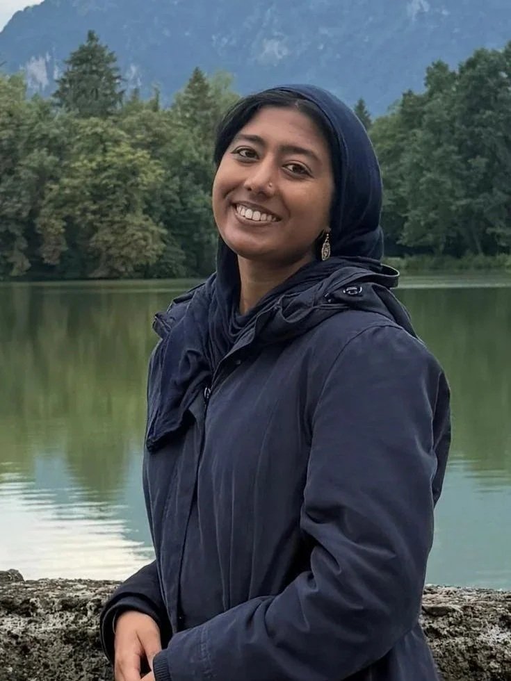 A woman in a dark blue jacket with a hood and scarf standing outdoors near a body of water, with trees and mountains in the background, smiling at the camera.