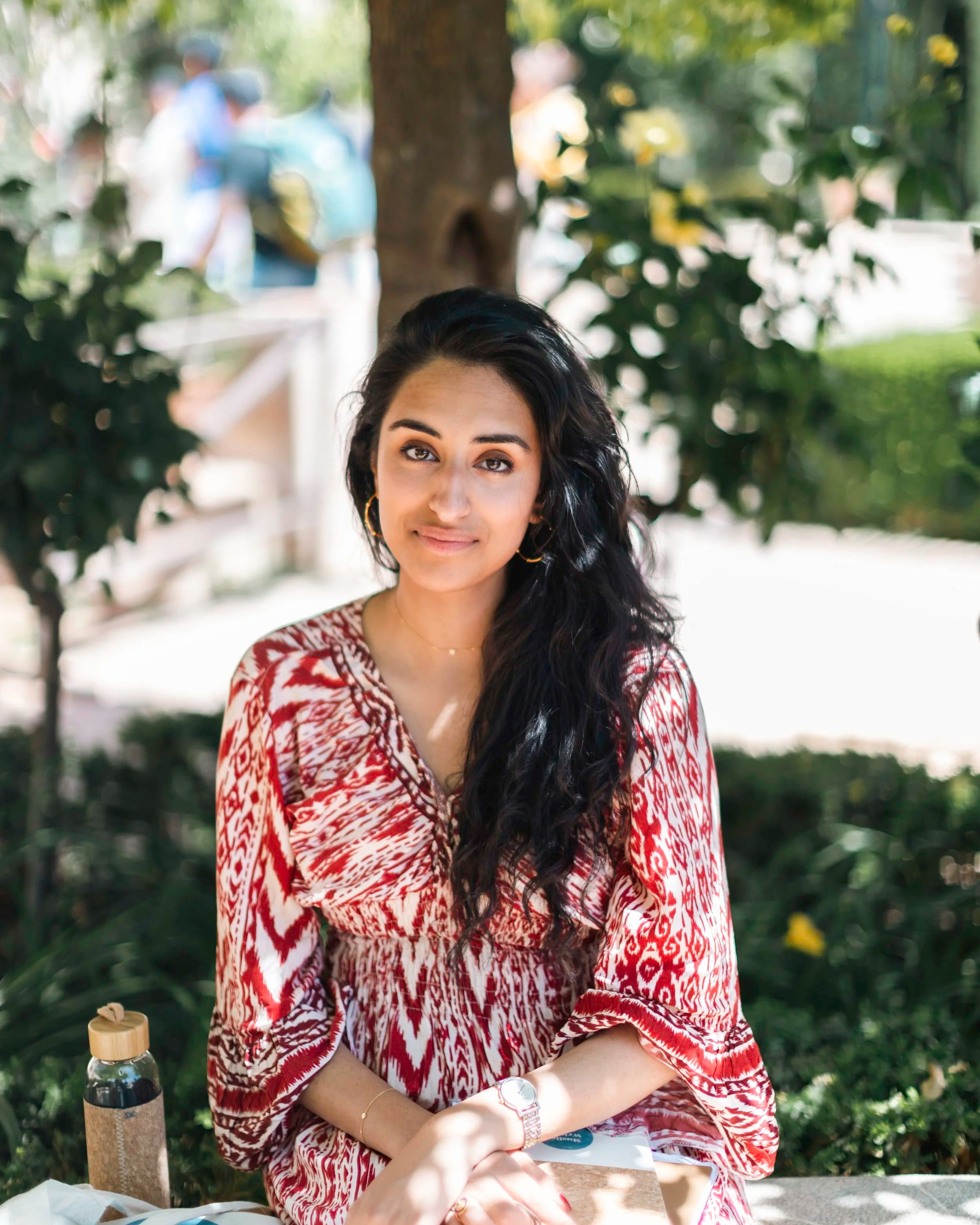 Young woman with long black curly hair wearing a red and white patterned dress, sitting outdoors at a table, smiling with greenery in the background.