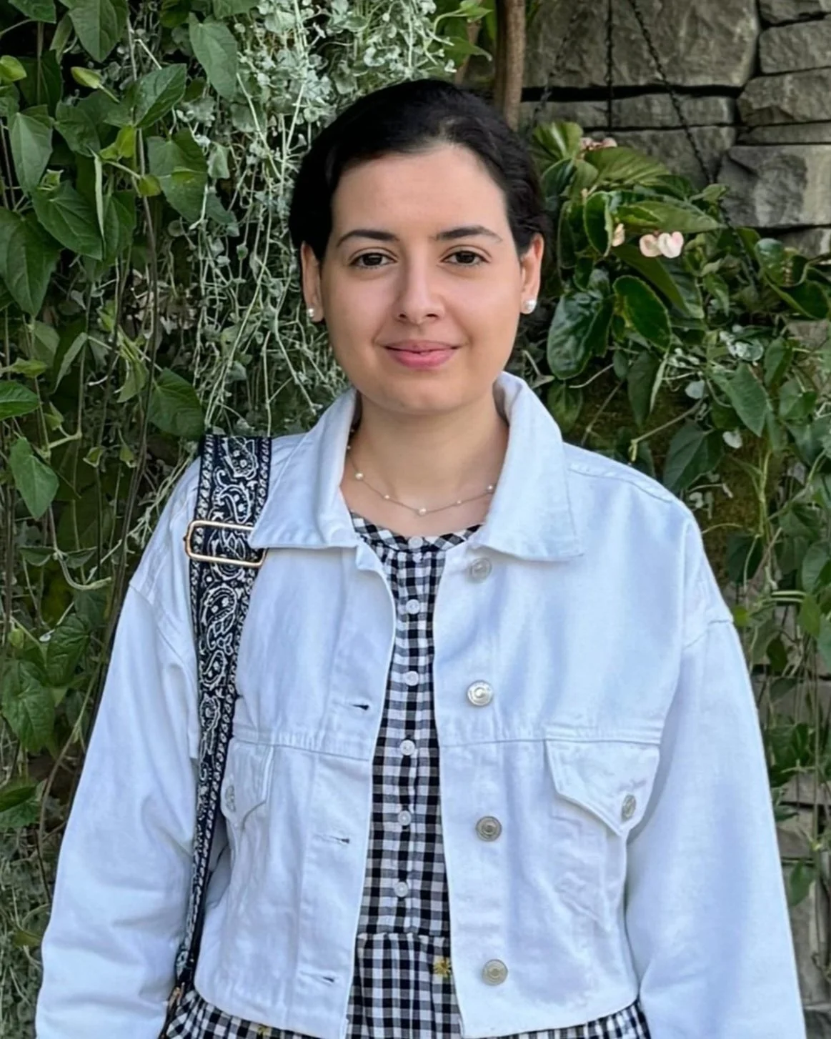 A woman with short dark hair wearing a white denim jacket, black and white checkered dress, pearl earrings, and a delicate necklace, standing in front of green foliage and a stone wall.