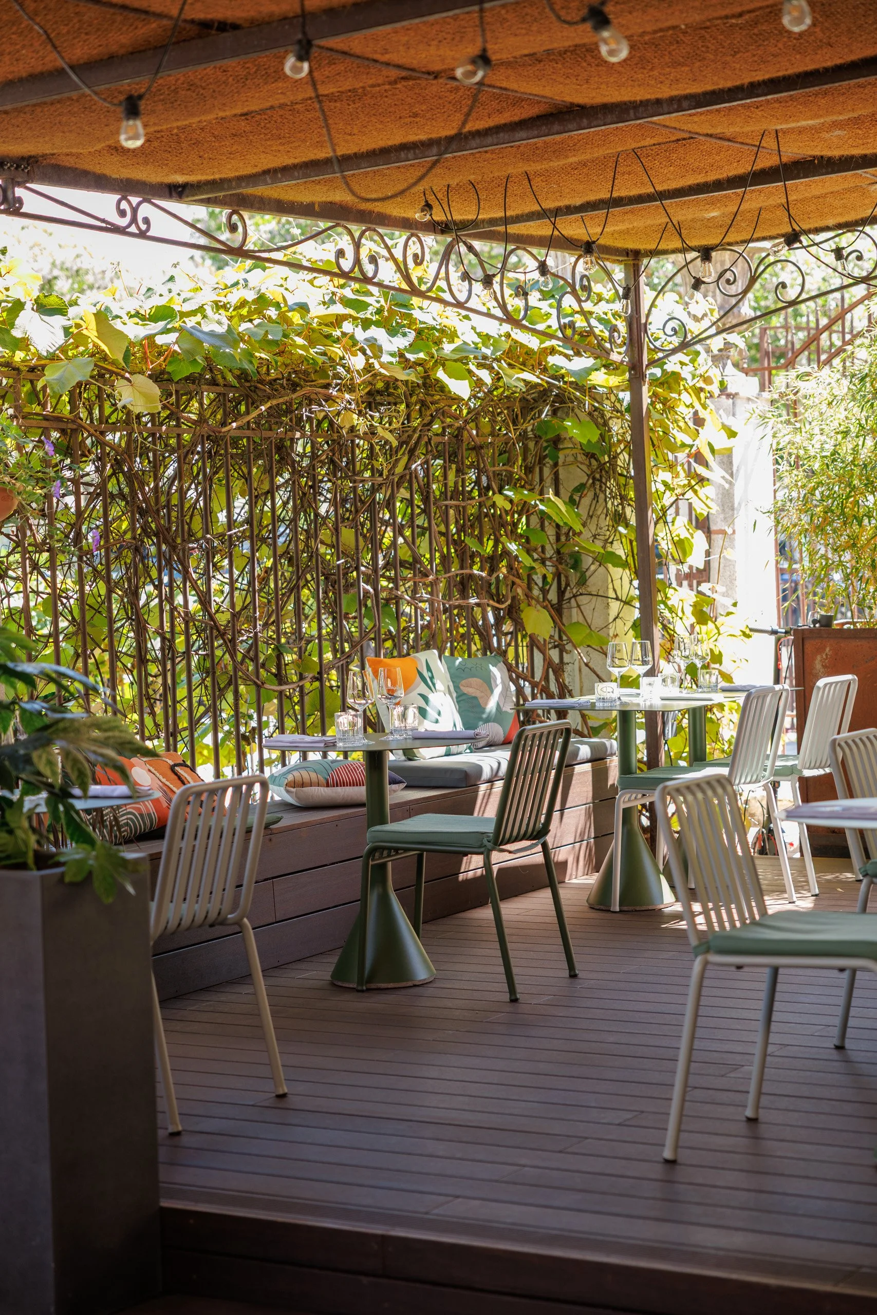 Une terrasse d'un restaurant ou café avec des tables, des chaises et plusieurs verres de vin, entourée de végétation et couverte partiellement par un toit en bois avec des guirlandes lumineuses.