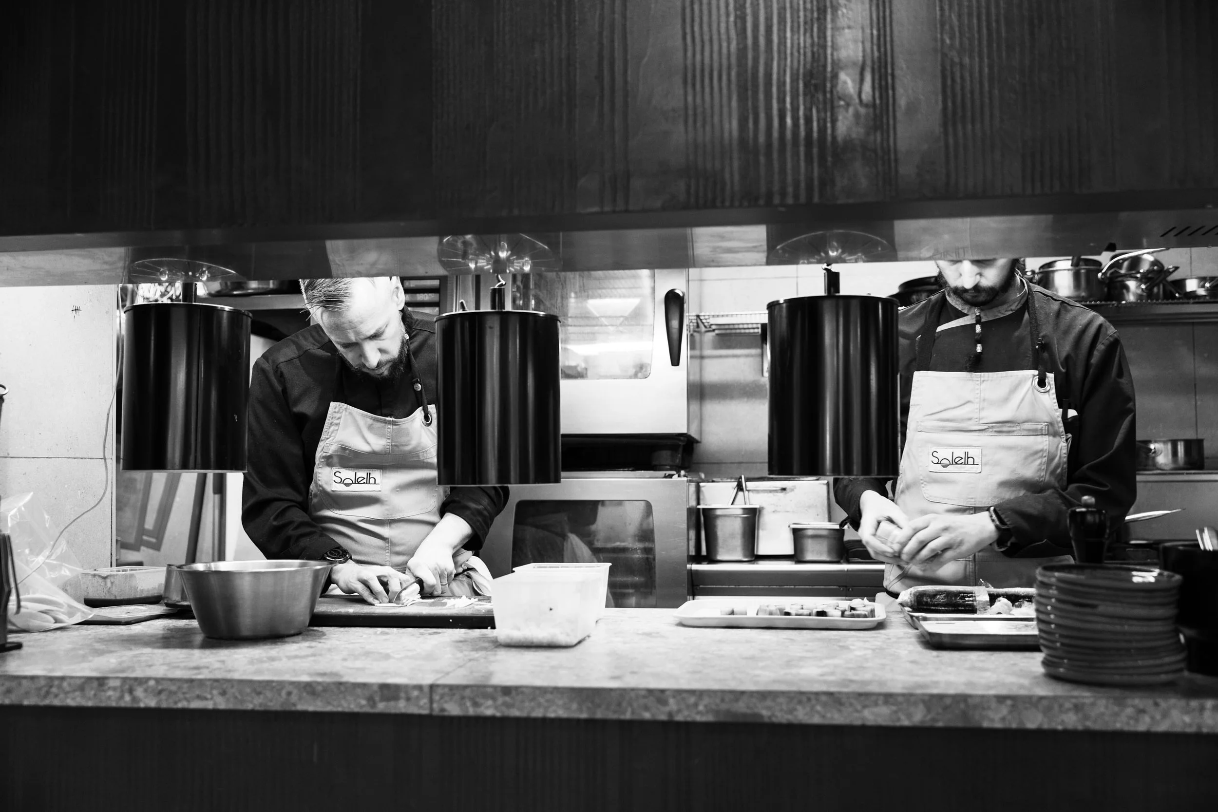 Deux chefs en cuisine, portant des tabliers, travaillent à une station de préparation. Vue partielle de leurs visages, concentrés sur leur tâche. Cuisine professionnelle avec ustensiles et équipements en arrière-plan.