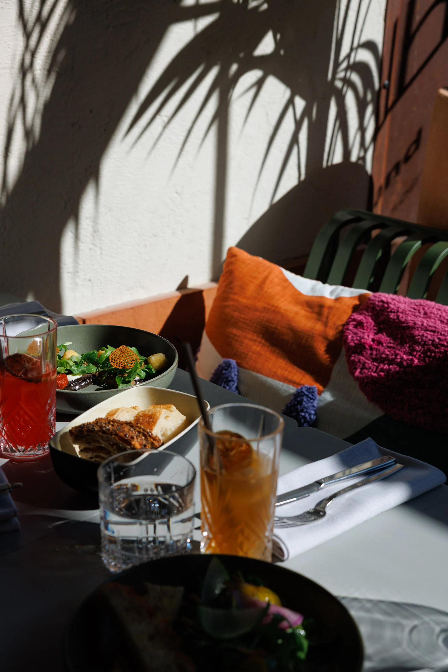 Une table de petit-déjeuner avec des plats, des boissons et des ustensiles, décorée avec des coussins colorés sur un banc à l'extérieur, avec des ombres de feuilles de palmier sur le mur en arrière-plan.