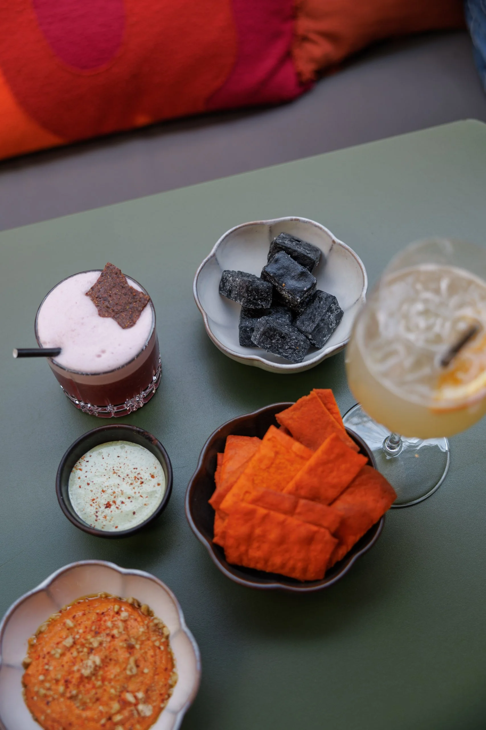 Assortiment de snacks et boissons, incluant un verre de boisson rouge avec une tranche de biscuit croustillant, un verre de boisson jaune avec citron, un bol de carrés de charbon noir, et divers dips et chips colorés sur une table.
