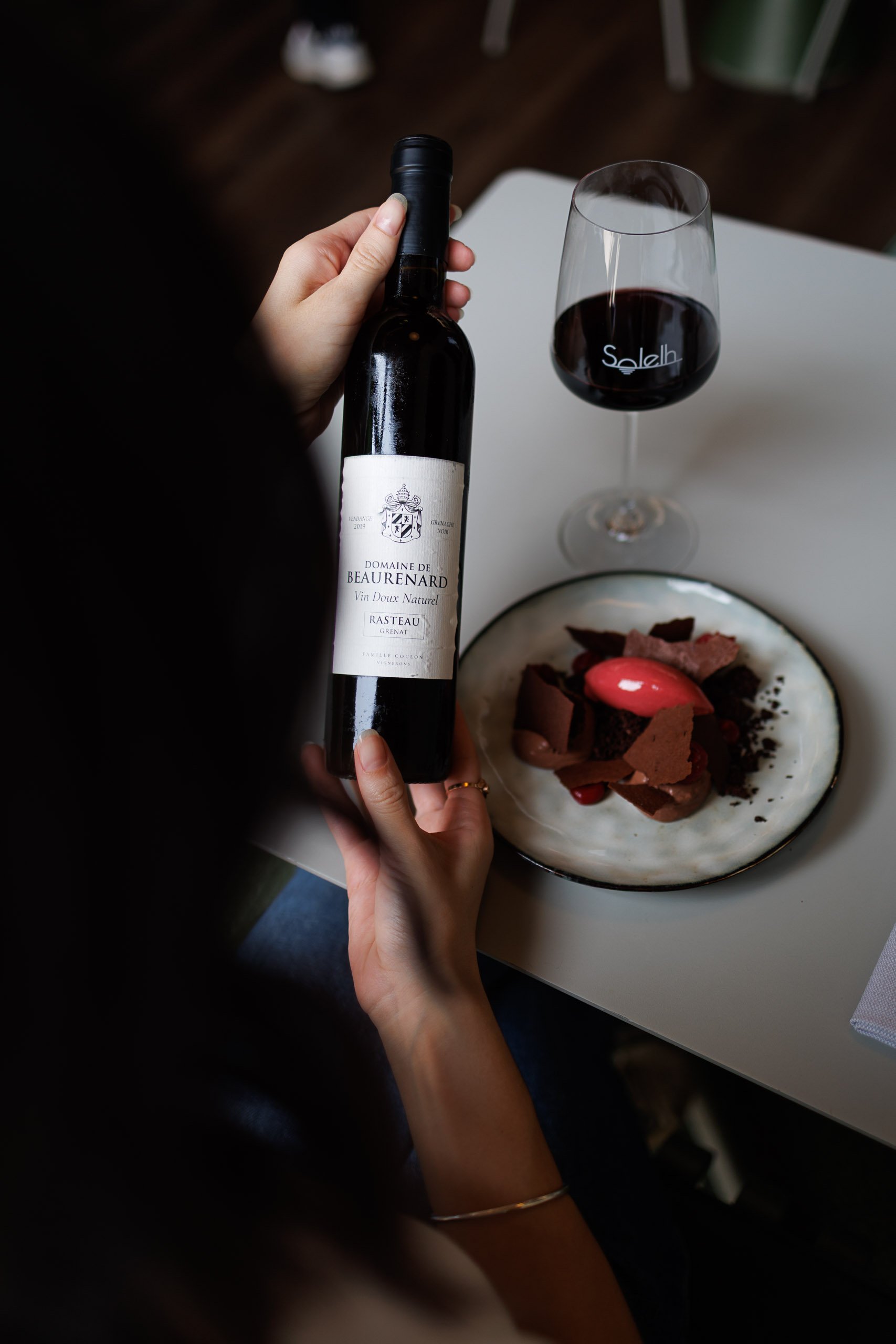 Une personne tient une bouteille de vin rouge Domaine de Beaurenard, et un verre de vin rempli de vin rouge est placé sur une table. Il y a aussi un dessert sur une assiette blanc crème à côté.