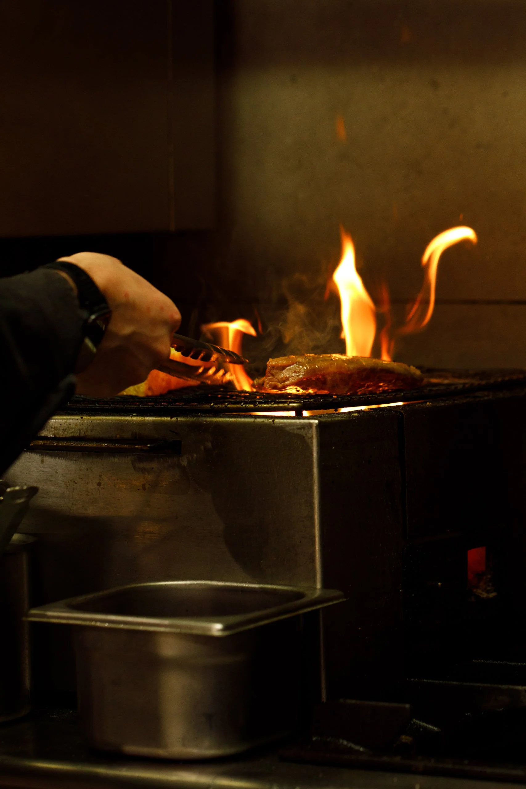 Une casserole en acier inoxydable sur une cuisinière avec des flammes de cuisson visibles, une personne utilisant une pince pour manipuler quelque chose à l'intérieur.