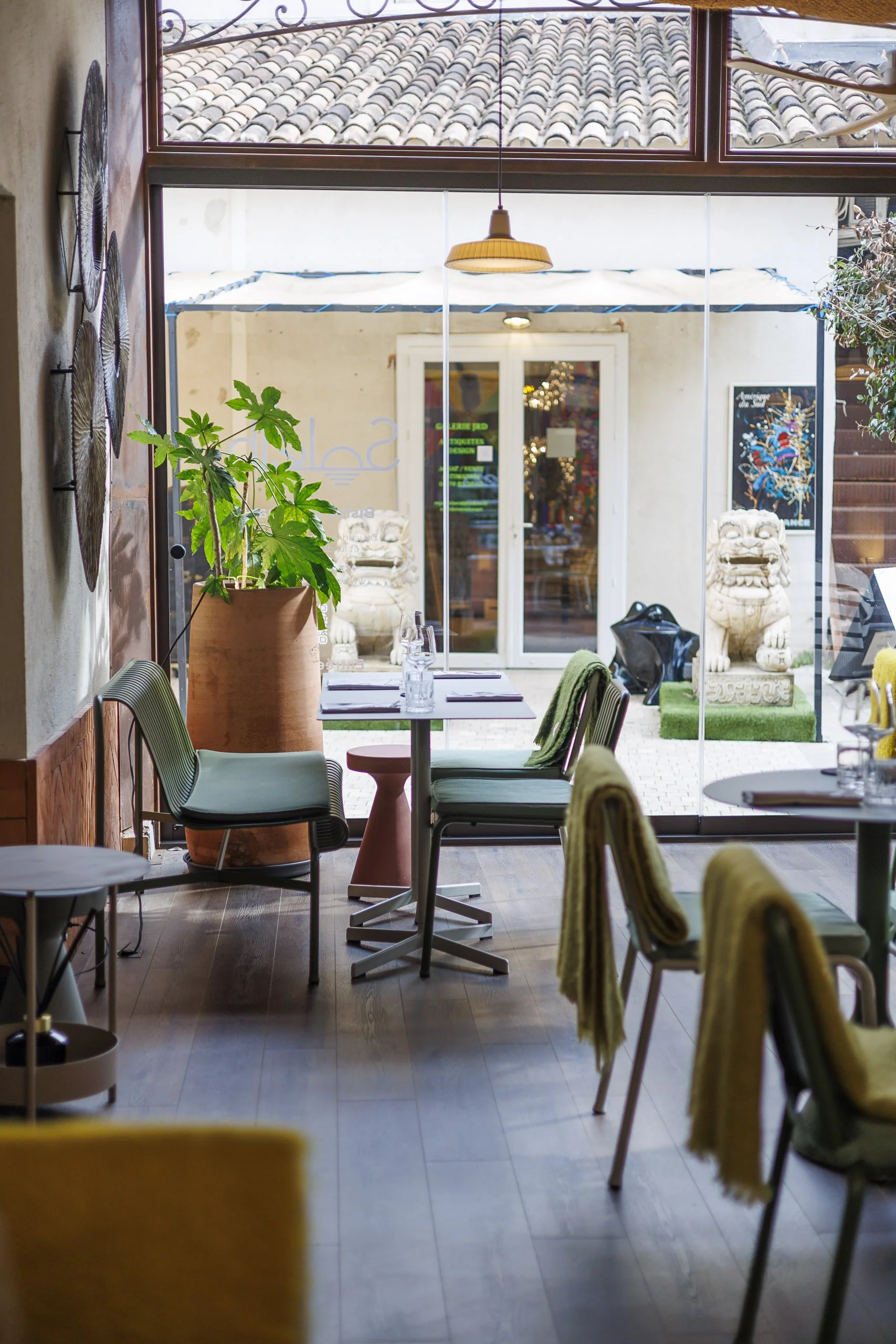 Intérieur d'un restaurant avec des tables, des chaises, des plantes vertes, une décoration murale en forme de disques et une vue sur une cour extérieure ornée de statues de lions en pierre et d'une porte en verre.