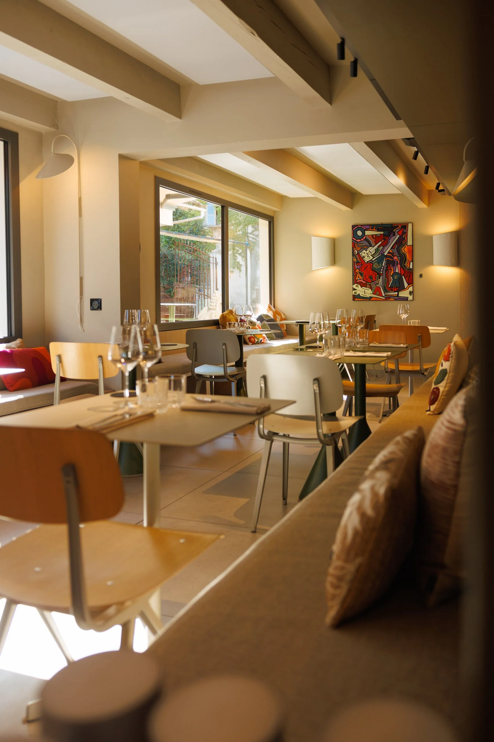 Intérieur d'un restaurant lumineux avec des fenêtres donnant sur un extérieur verdoyant, décor moderne avec tableau coloré sur le mur et tables dressées avec verres et couverts.