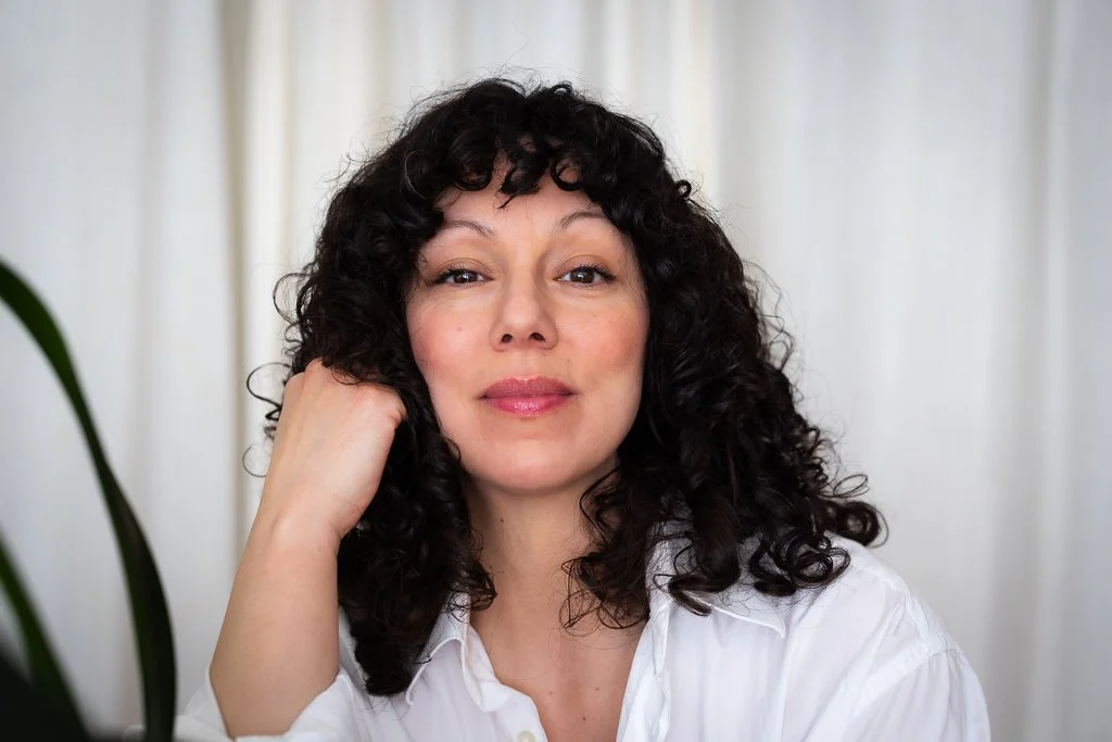 Femme avec des cheveux bouclés noirs, portant une chemise blanche, assise devant un fond clair avec une plante verte partielle visible à gauche.