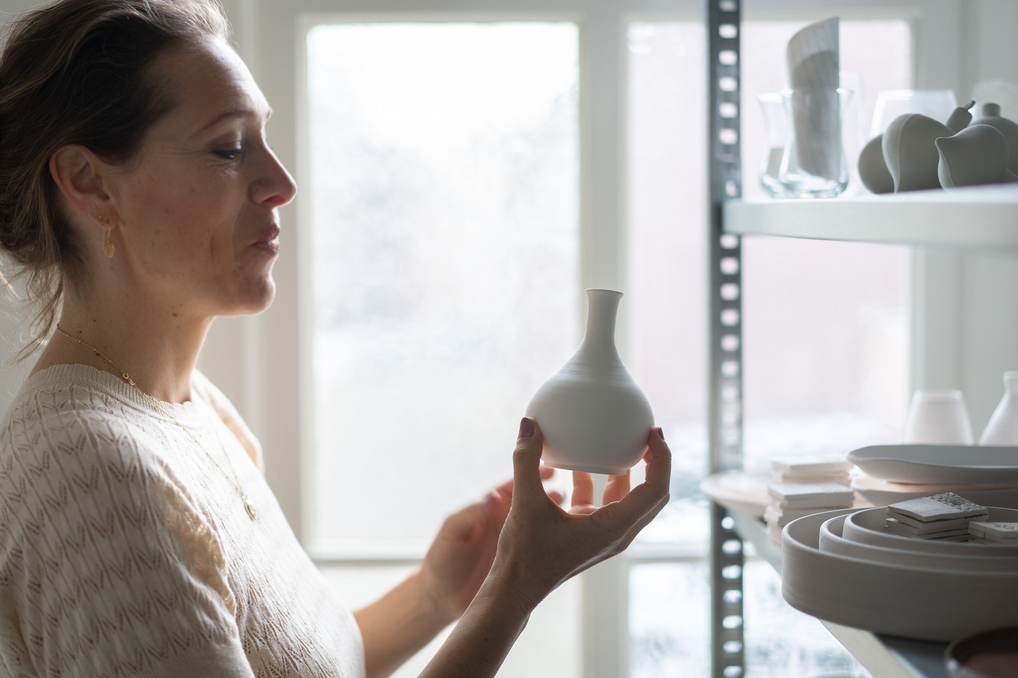 Vrouw houdt witte vaas vast in lichte kamer met planken vol keramiek.