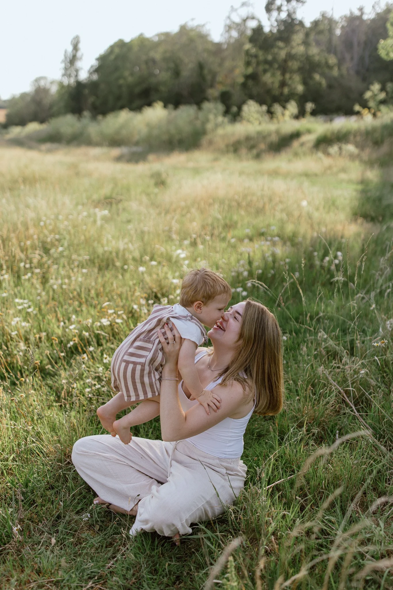 A woman with long brown hair, wearing a white tank top and cream pants, sitting on the grass in a field of wildflowers, lifting a young boy with red hair and a striped shirt into the air. They are smiling and looking at each other, with trees and a s