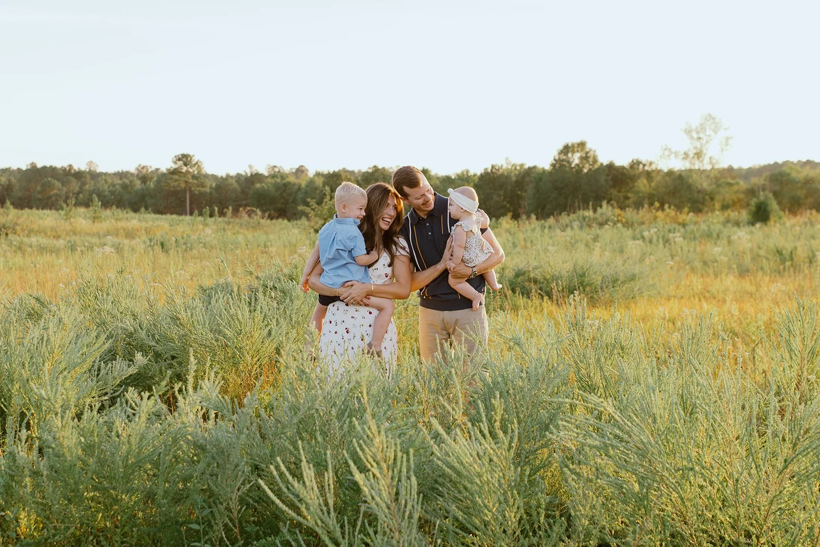 Family of four enjoying a walk through a green field at sunset, with trees in the background.