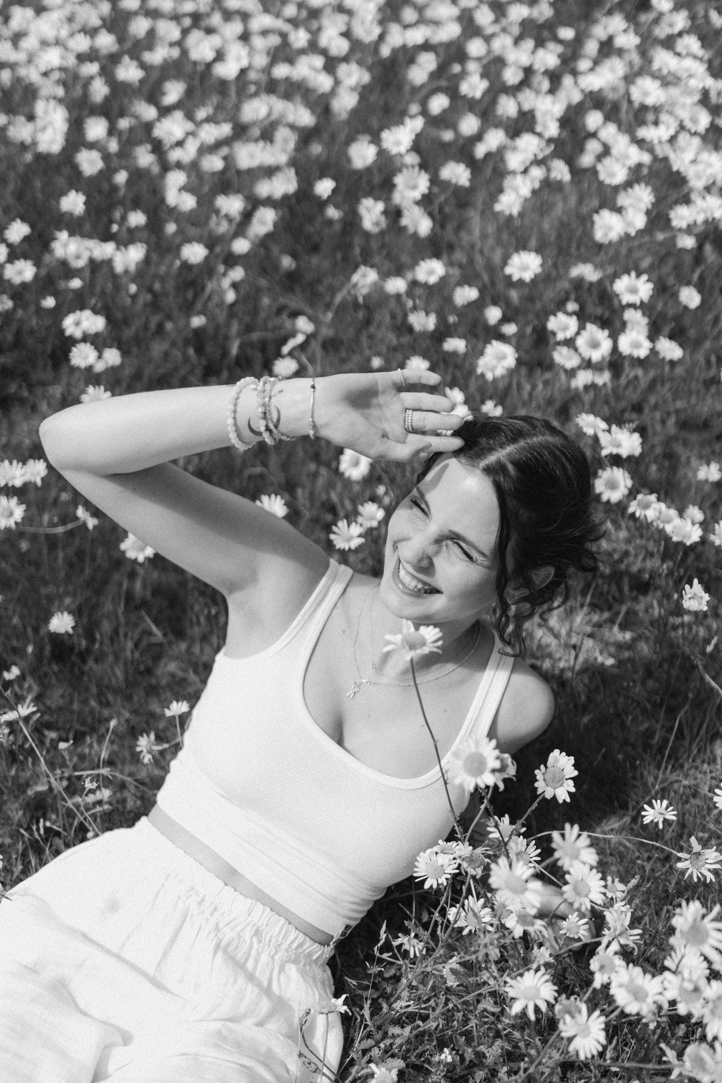 A smiling woman lying in a field of daisies, shielding her eyes from the sun with one hand. She wears a white tank top, loose white pants, bracelets, rings, and a delicate necklace, with daisies surrounding her. The photograph is in black and white.