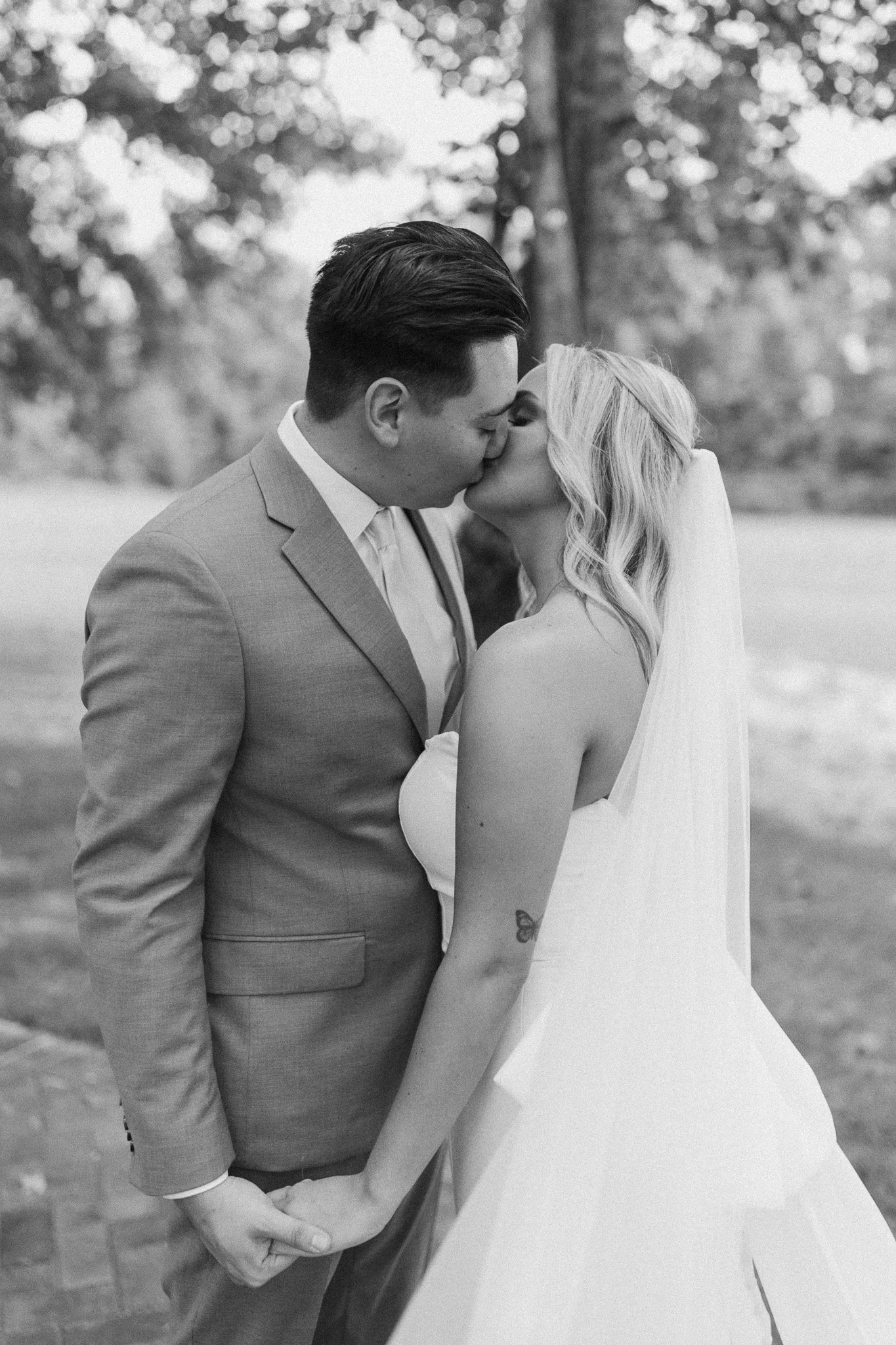 A black-and-white photograph of a couple in wedding attire, kissing outdoors. The groom is wearing a suit, and the bride is in a white wedding dress and veil. They are holding hands with intertwined fingers. The background shows trees and blurred foliage.