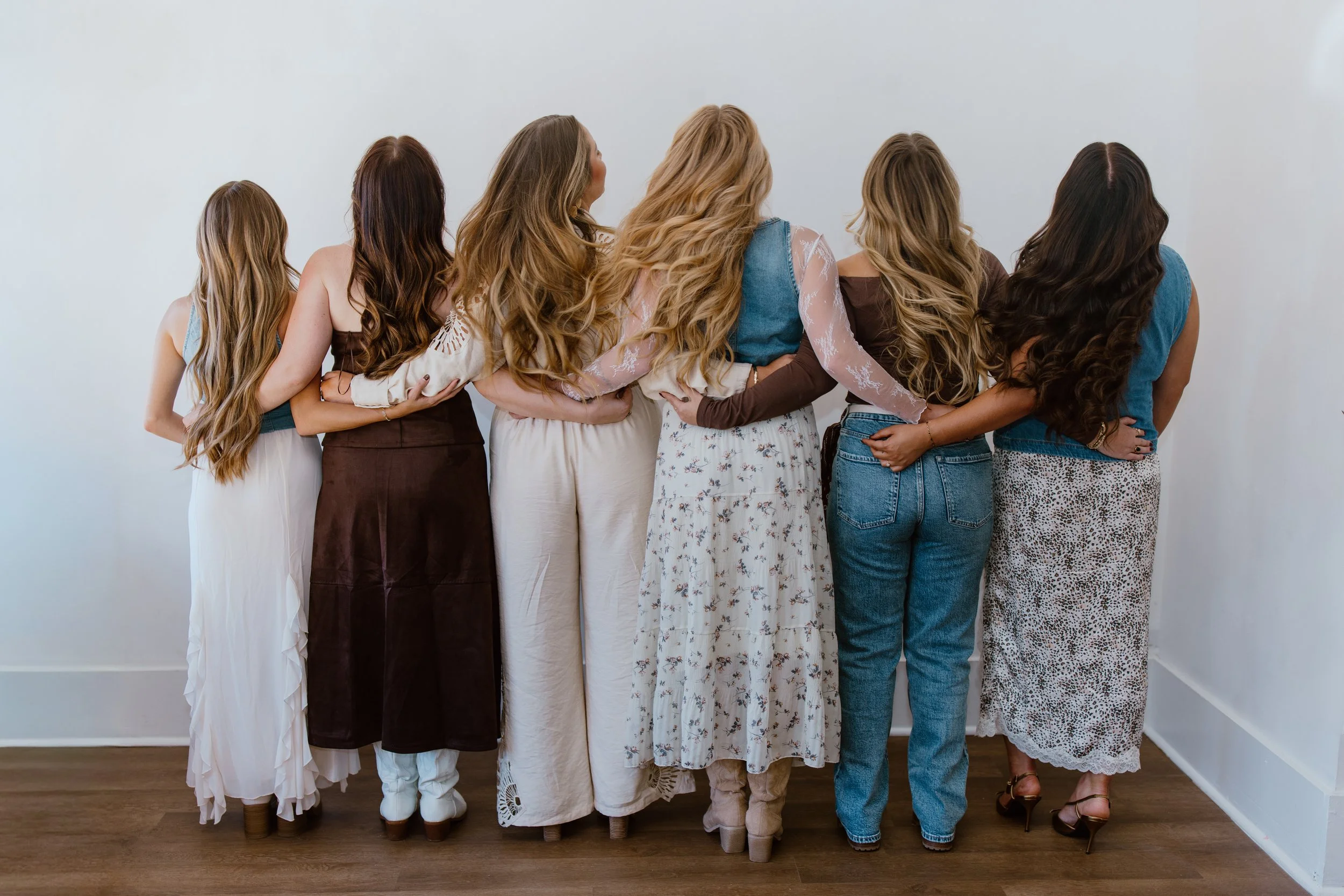 Six women with long hair standing with their backs to the camera, arms wrapped around each other's waists, against a plain white wall.