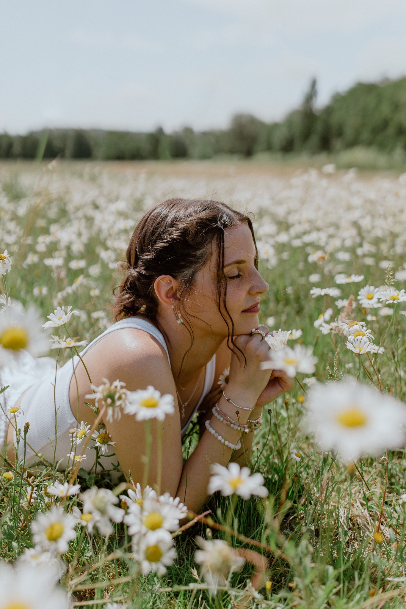 A woman with dark hair styled in a braid is lying in a field of white daisies with her eyes closed and a serene expression, surrounded by lush greenery and a bright sky.