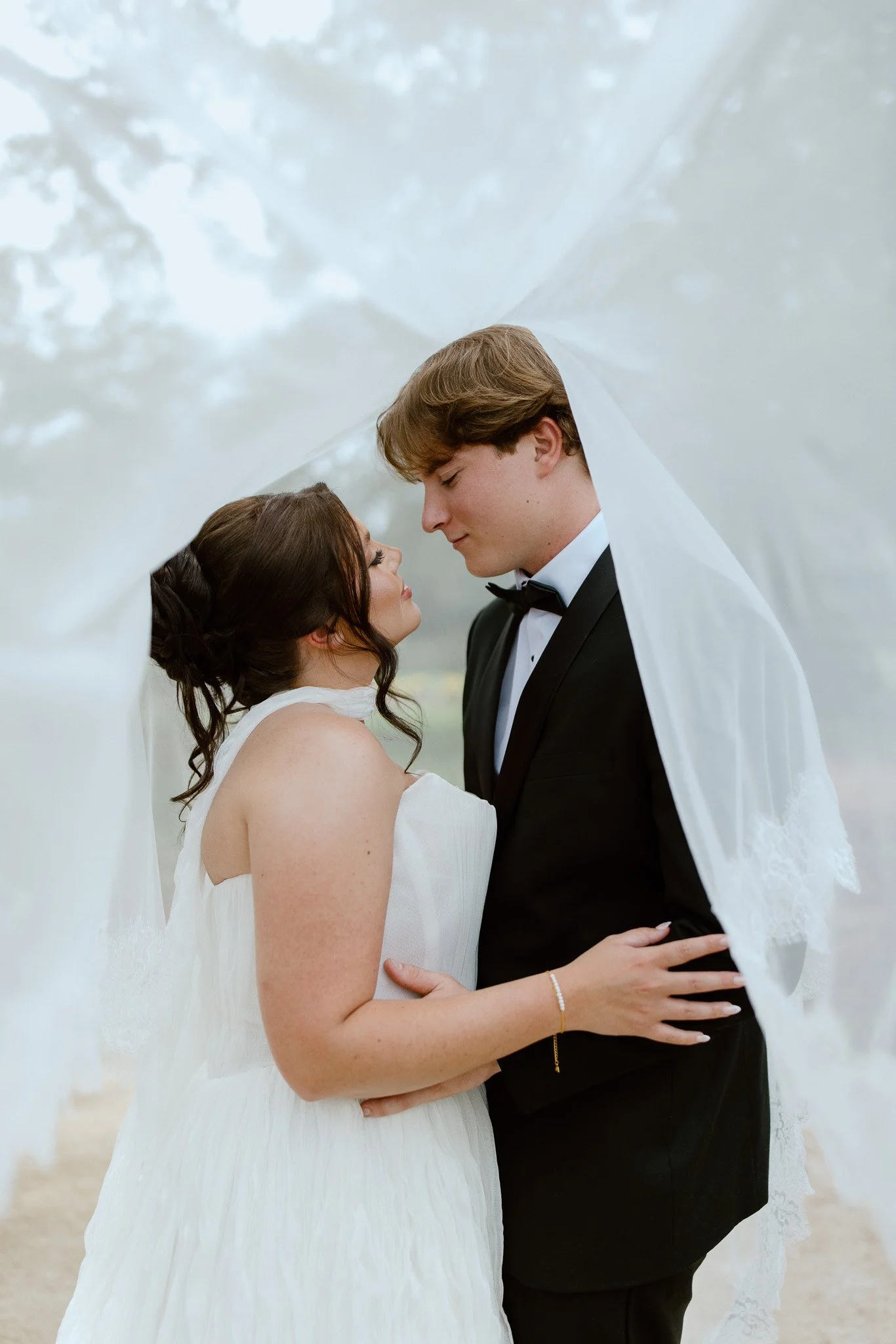 A bride and groom share a romantic moment, standing under a white wedding veil outdoors, with trees and a soft sky in the background.