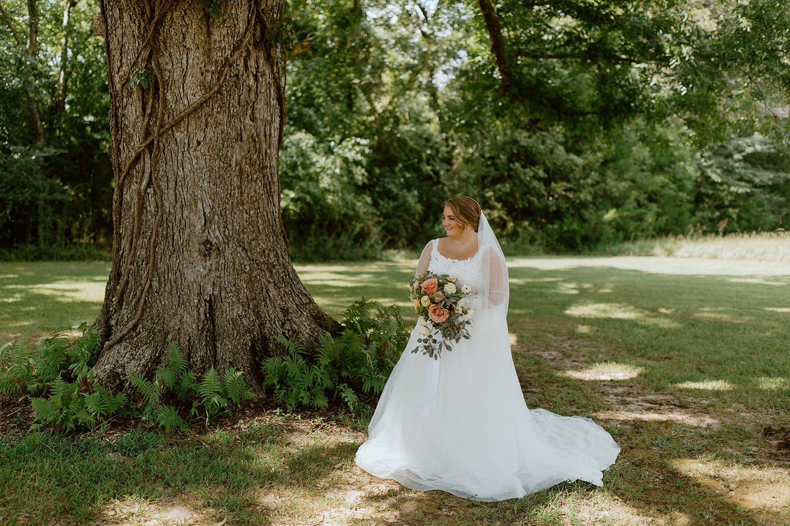A bride in a white wedding dress holding a bouquet of flowers, standing outdoors next to a large tree with green foliage in the background.