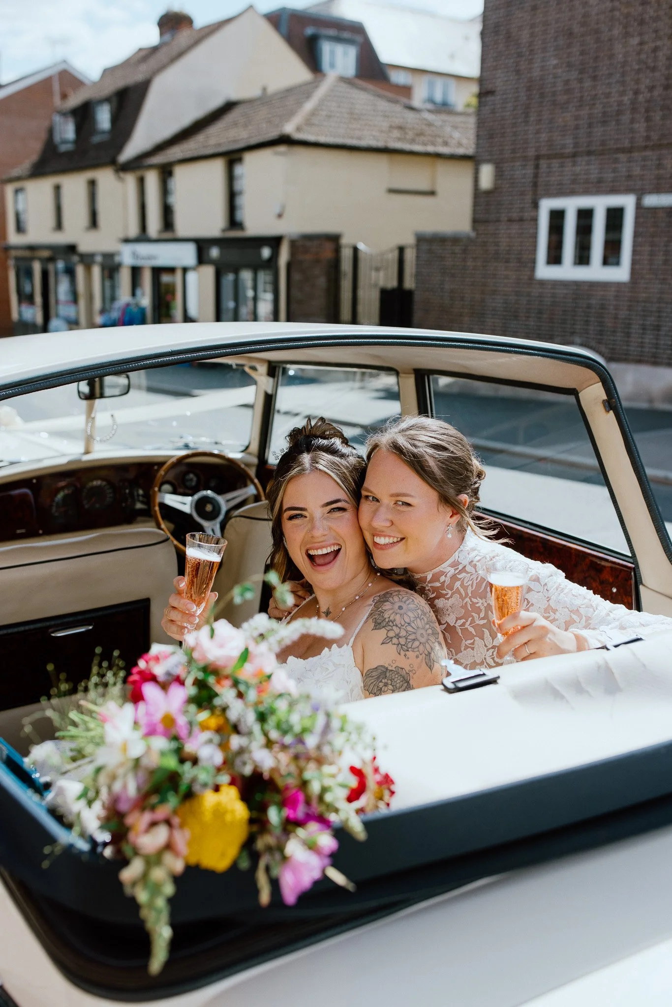 Two smiling women dressed in wedding attire sitting inside a vintage convertible car, holding glasses of champagne, with a colorful flower bouquet on the front of the car and a street with houses in the background.