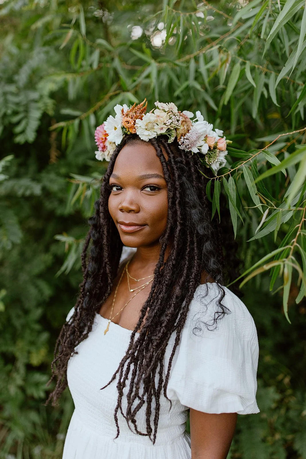 A woman with dark skin and long curly hair wearing a white dress and layered gold necklaces, standing outdoors among green foliage, with a flower crown on her head.