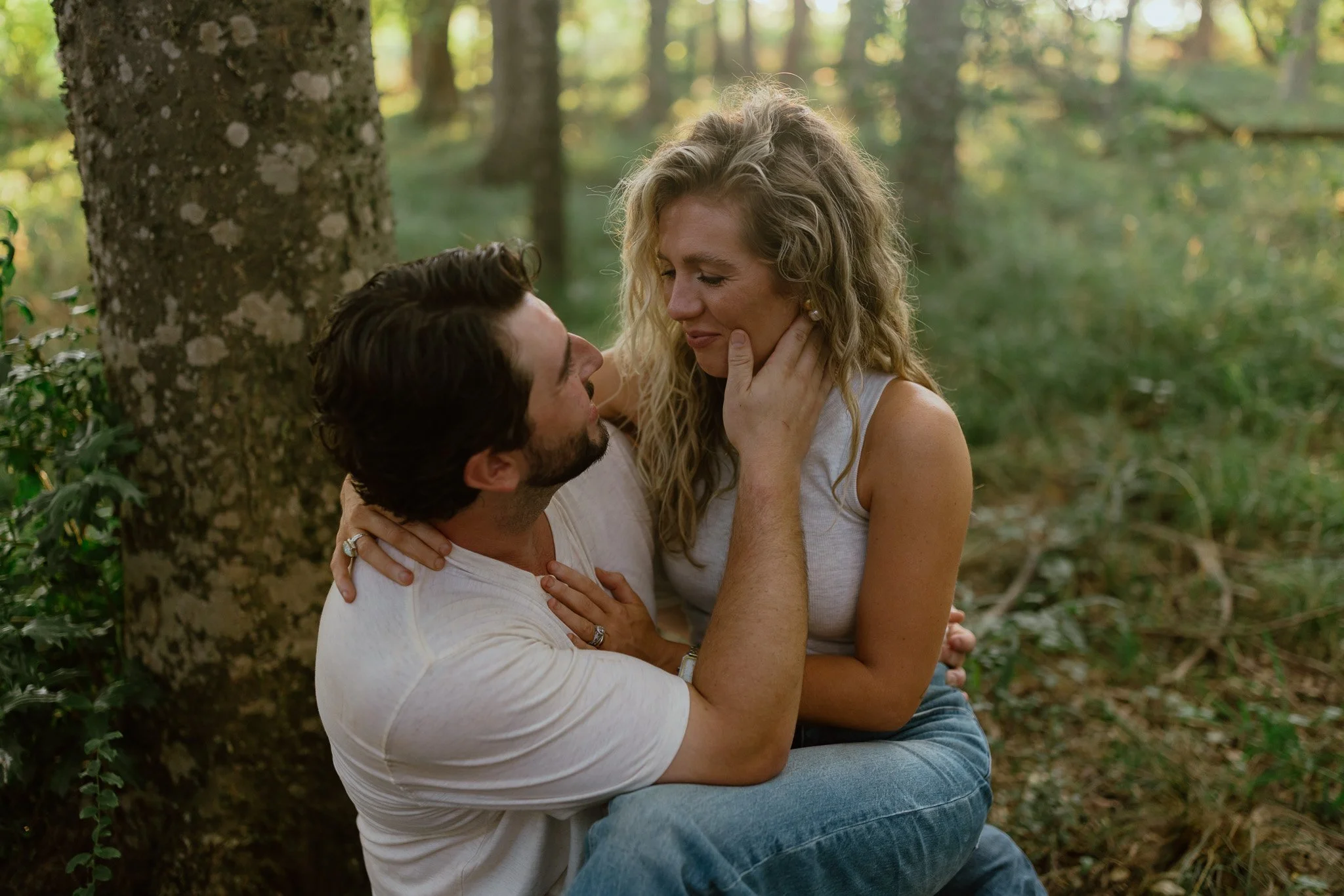 A man and woman sitting close together outdoors in a wooded area, with the man holding the woman's face tenderly and both smiling emotionally.