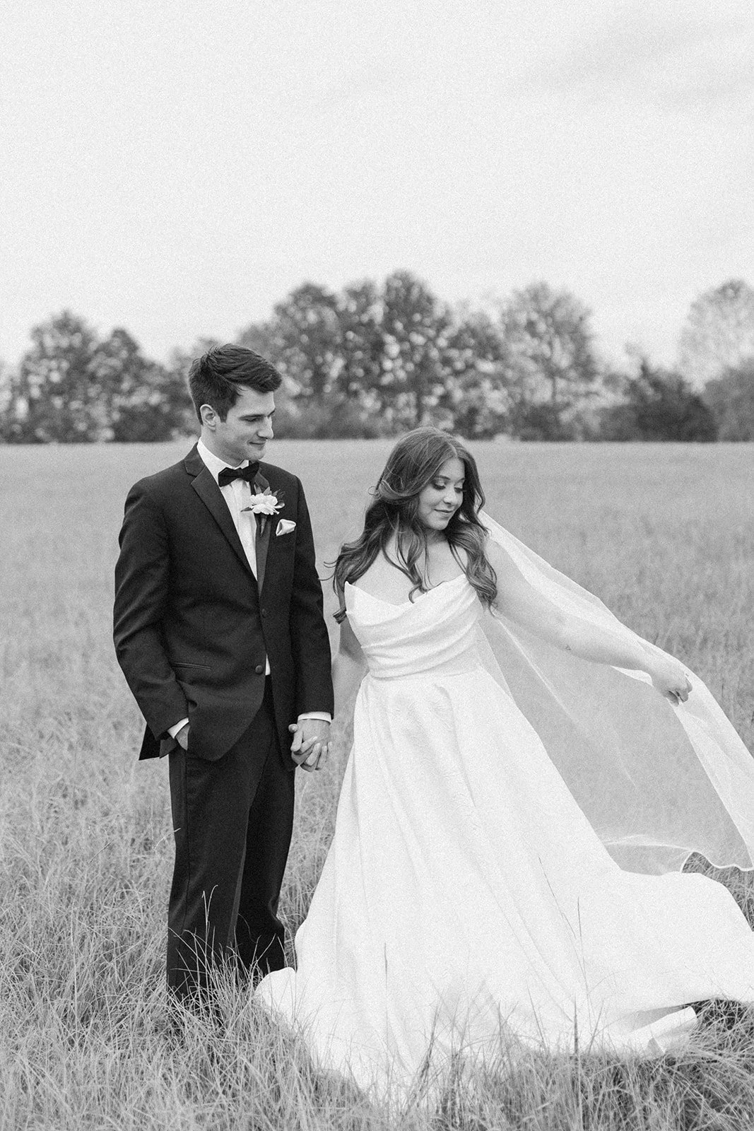 Black-and-white portrait of bride and groom walking through a field