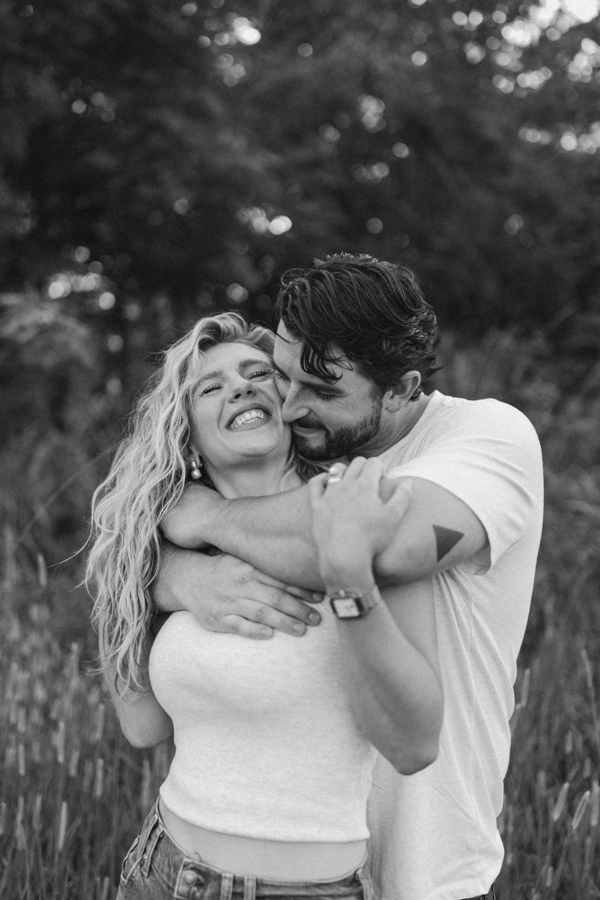 A black and white photo of a happy couple hugging outdoors, with the woman smiling and the man leaning in close, surrounded by trees.