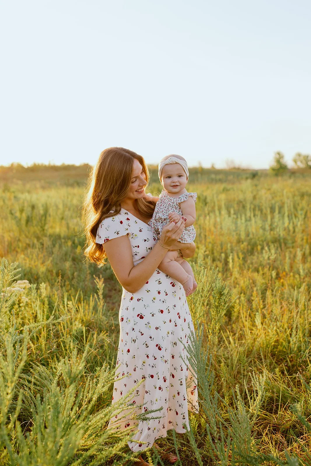 A woman holding a baby girl in a grassy field during sunset, both smiling, woman wearing a white floral dress, baby with headband and floral dress.