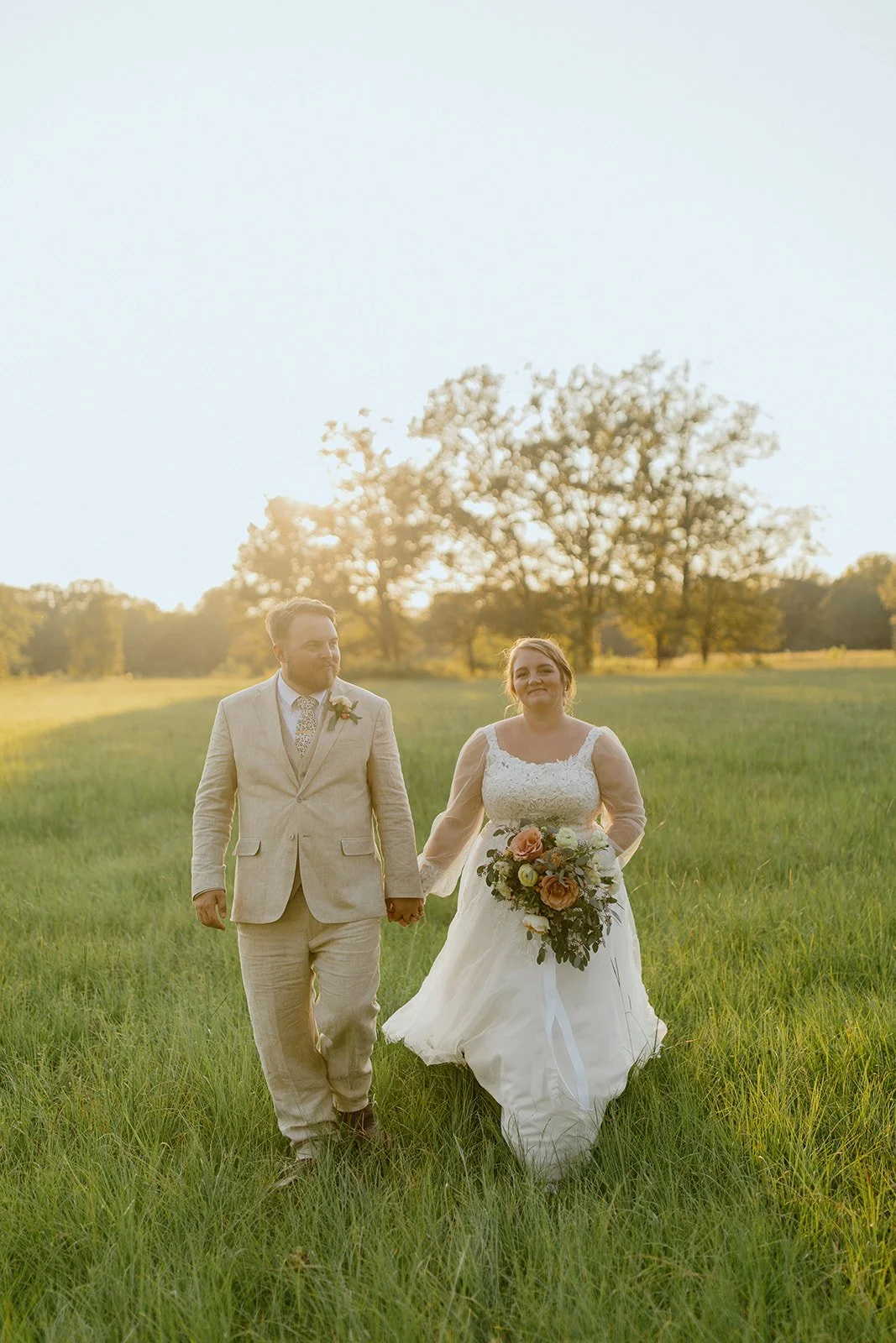 A bride and groom holding hands walking through a grassy field at sunset. The bride wears a white lace wedding gown and holds a large bouquet of flowers. The groom wears a beige suit with a patterned tie. Trees are visible in the background, and the sky is clear with the sun setting.