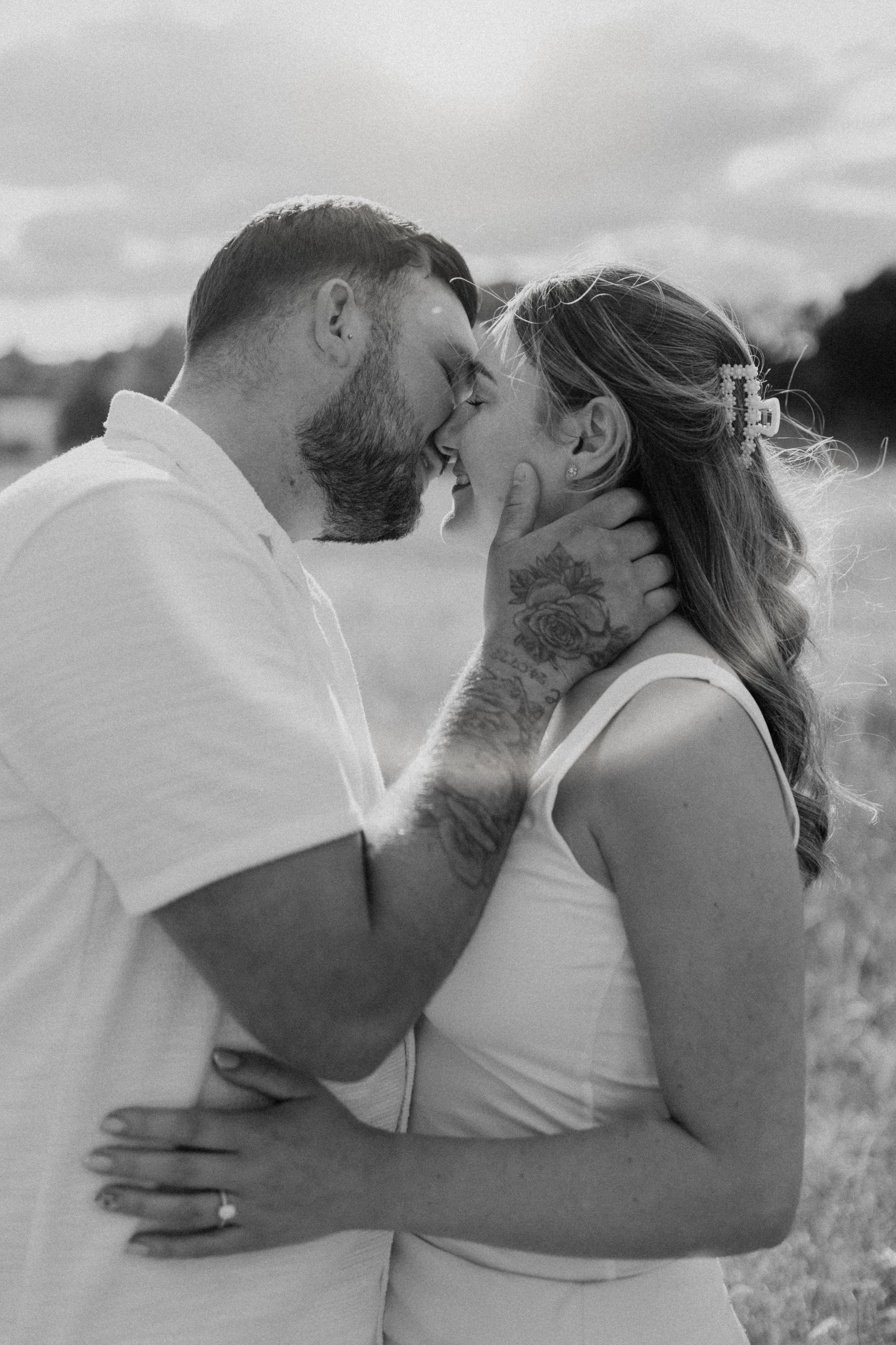Black and white photo of a couple touching foreheads and smiling, with the man gently holding the woman's face, outdoors with a blurred natural background.