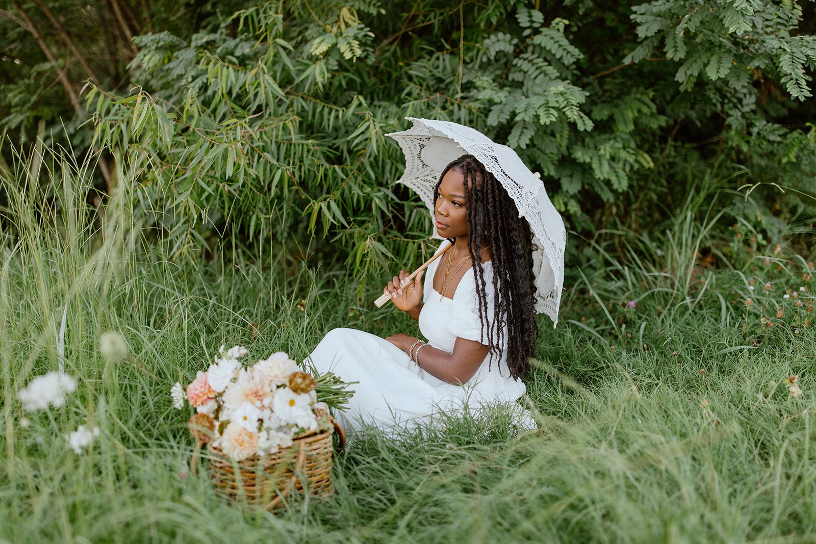 A woman in a white dress sitting in a grassy field holding a lace parasol with a basket of flowers nearby.