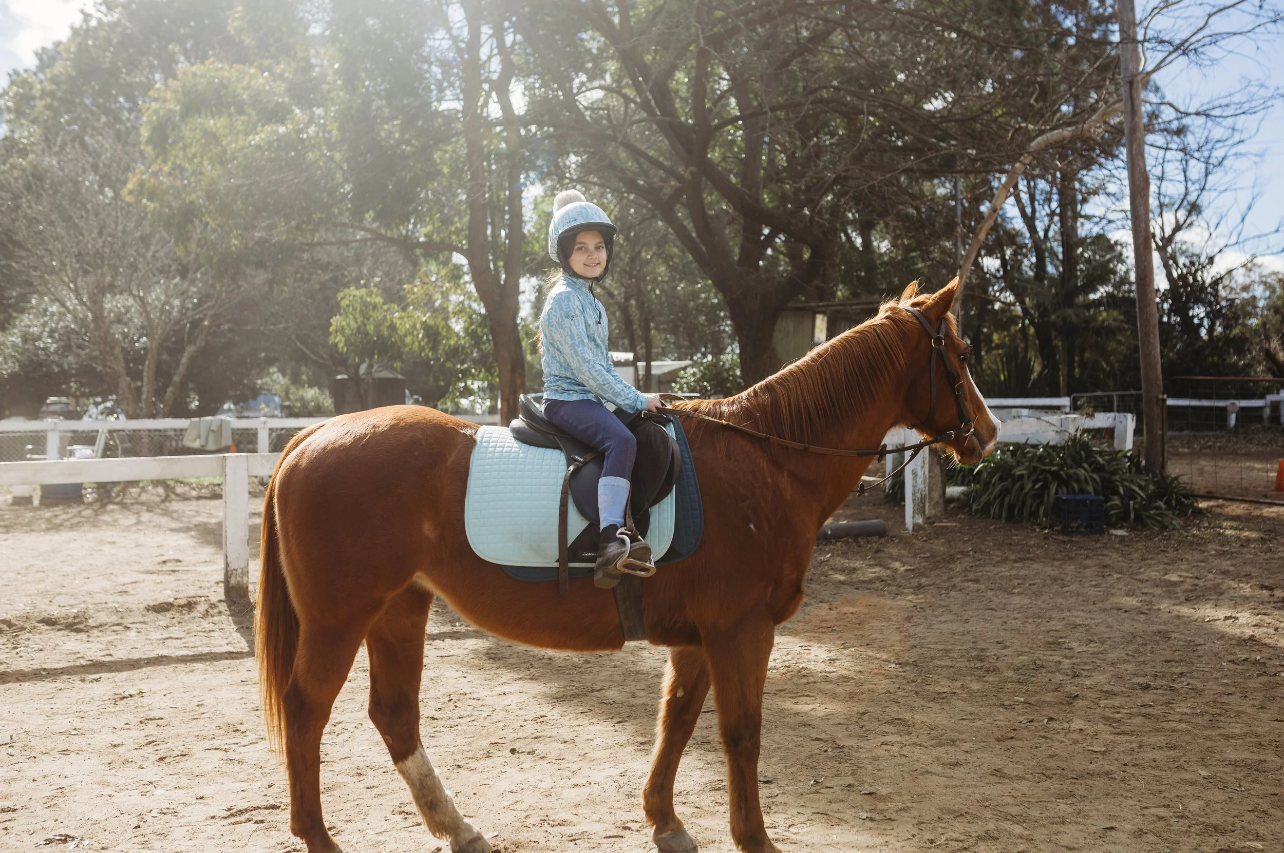 A young girl wearing a helmet and riding gear, sitting on a brown horse with a blue saddle pad in an outdoor riding arena surrounded by trees and fencing.