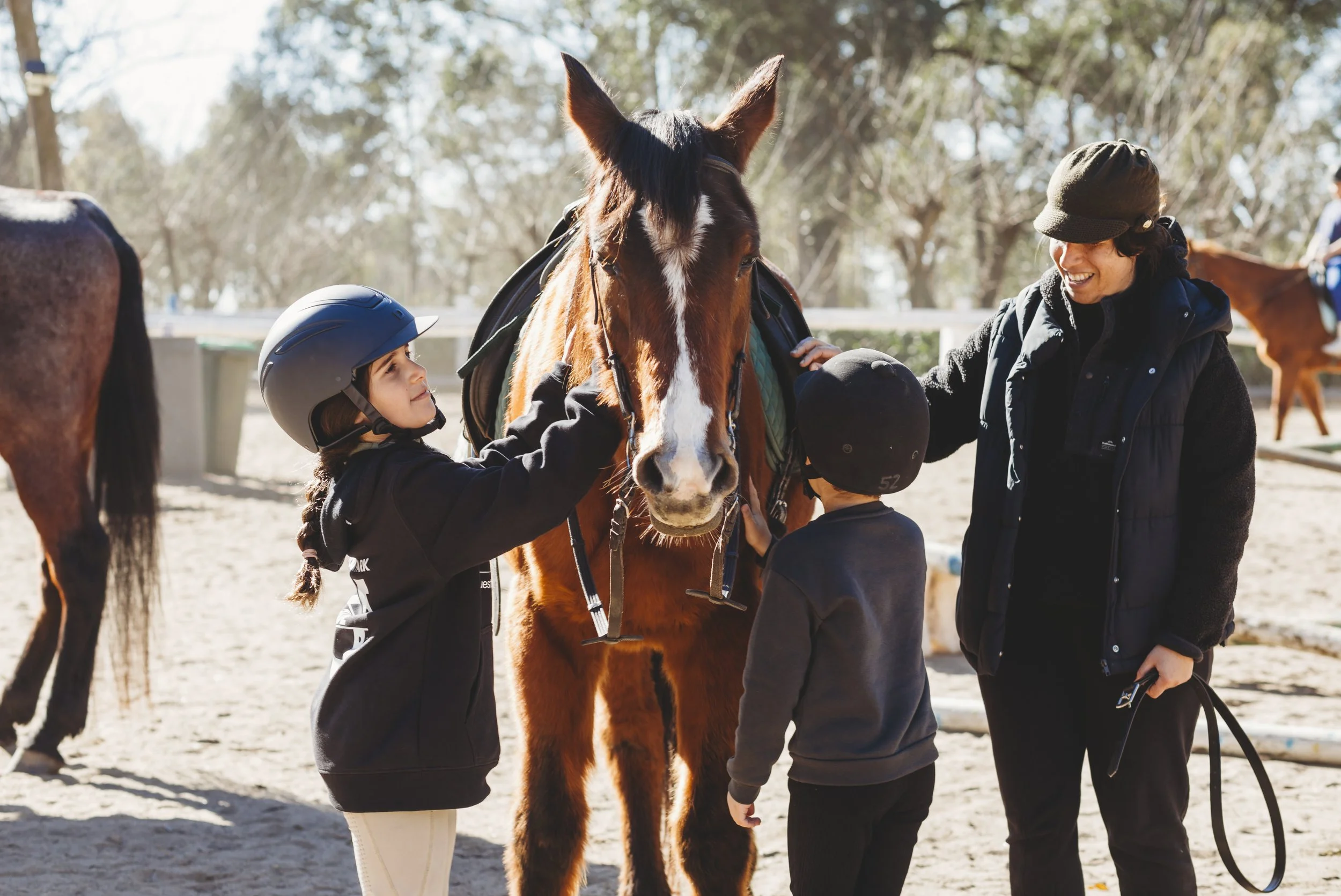 Two children and a woman with a brown horse outdoors on a sunny day, children wearing helmets, woman smiling, trees in background.