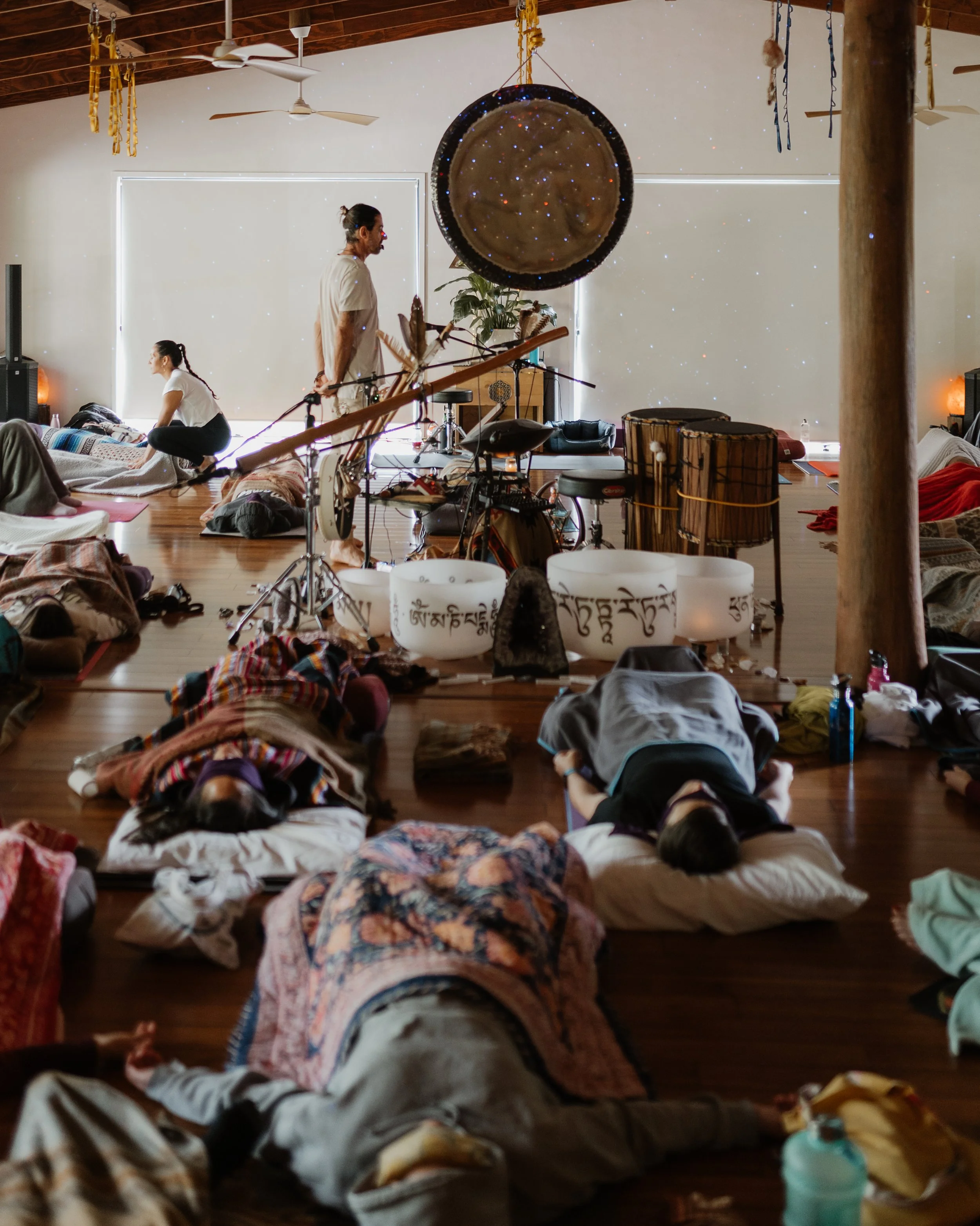 People practicing yoga in a spacious room with wooden floors, musical instruments, and large windows with blinds.