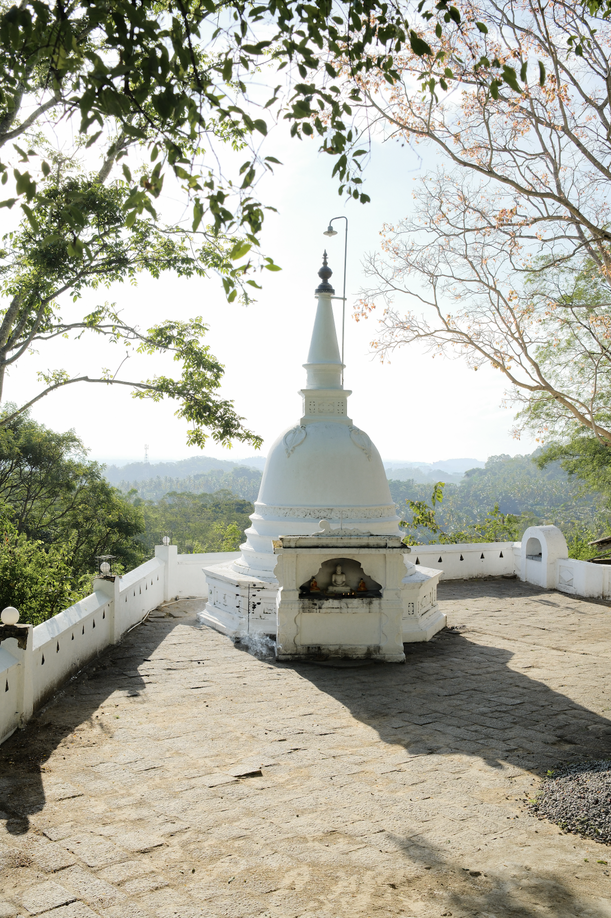 A white Buddhist stupa or pagoda situated on a stone platform, surrounded by trees with green and sparse pink leaves, under a bright sky.