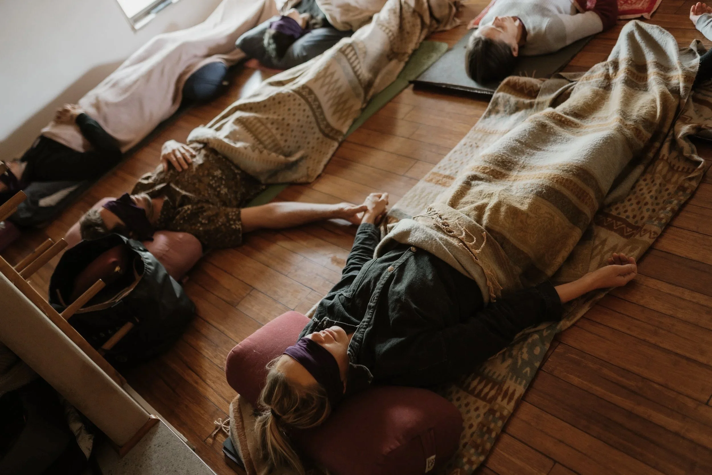 People lying on yoga mats and blankets with eyes closed, practicing meditation in a cozy room with wooden floors.