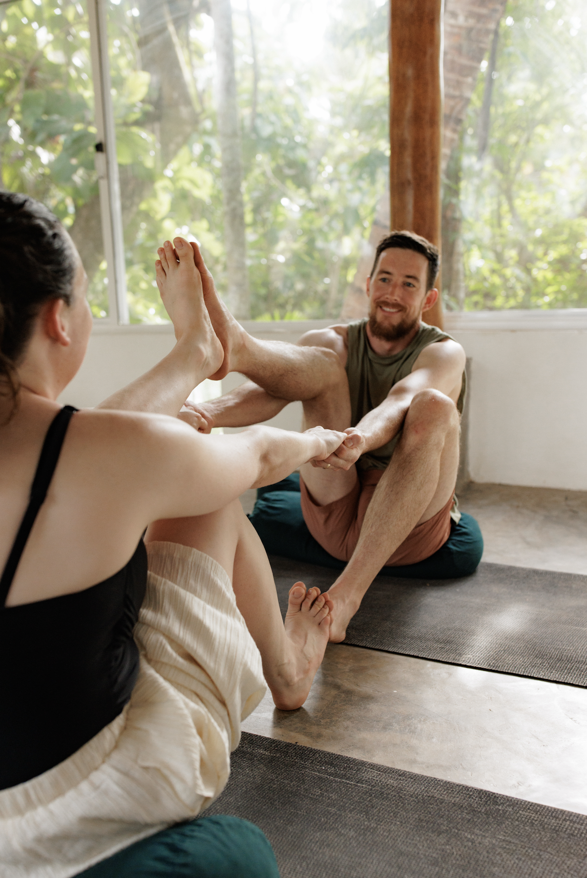 A man and woman practicing partner yoga indoors with large windows showing greenery outside. The woman is sitting with her legs crossed, reaching forward to hold the man's hands, who is sitting with his legs bent and his right arm extended, smiling at her.