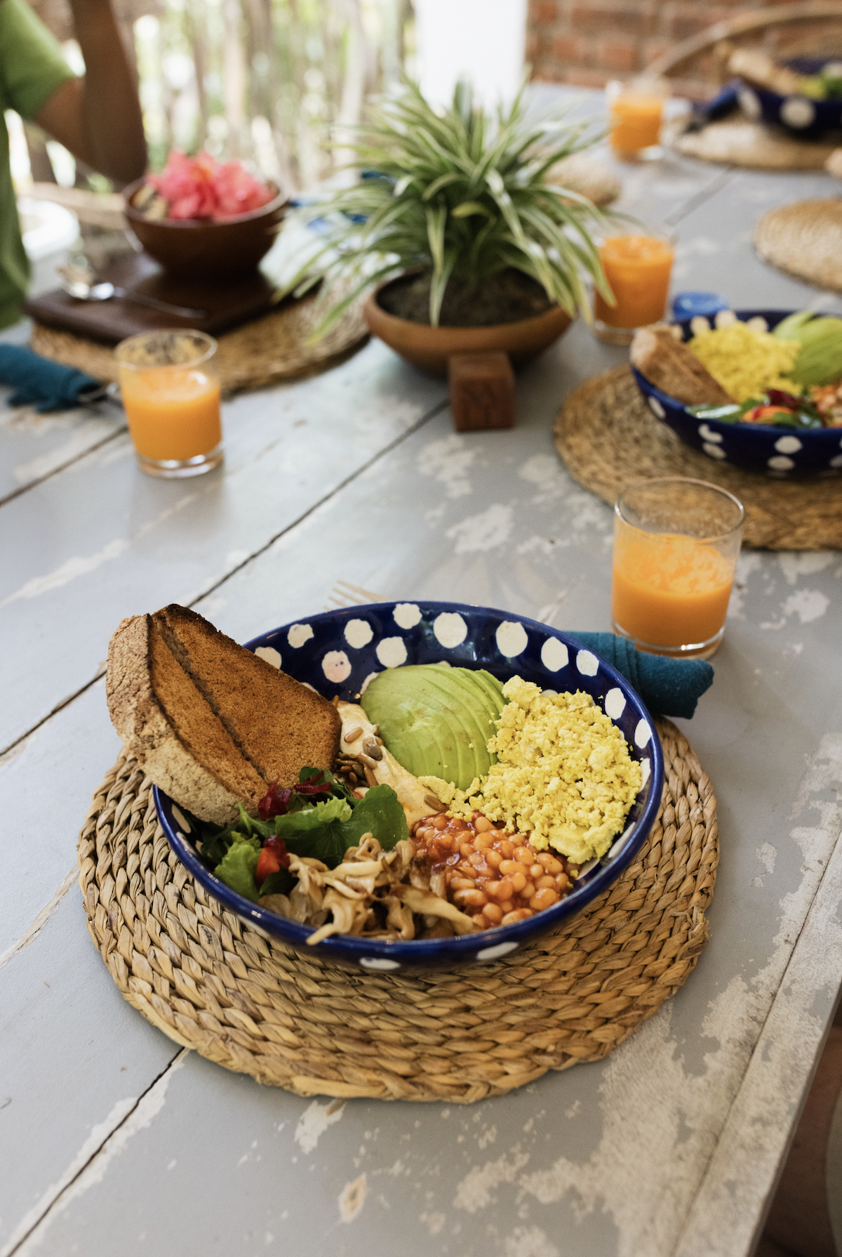 A colorful bowl of food on a woven placemat, containing sliced avocado, scrambled eggs, baked beans, shredded chicken, and slices of toasted bread.
