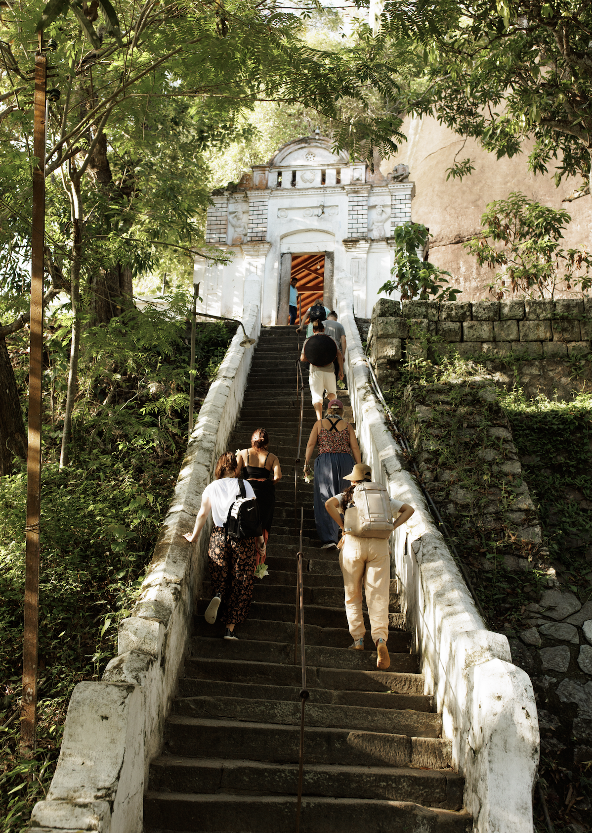 People climbing a long stone staircase towards an old white building entrance surrounded by lush green trees.