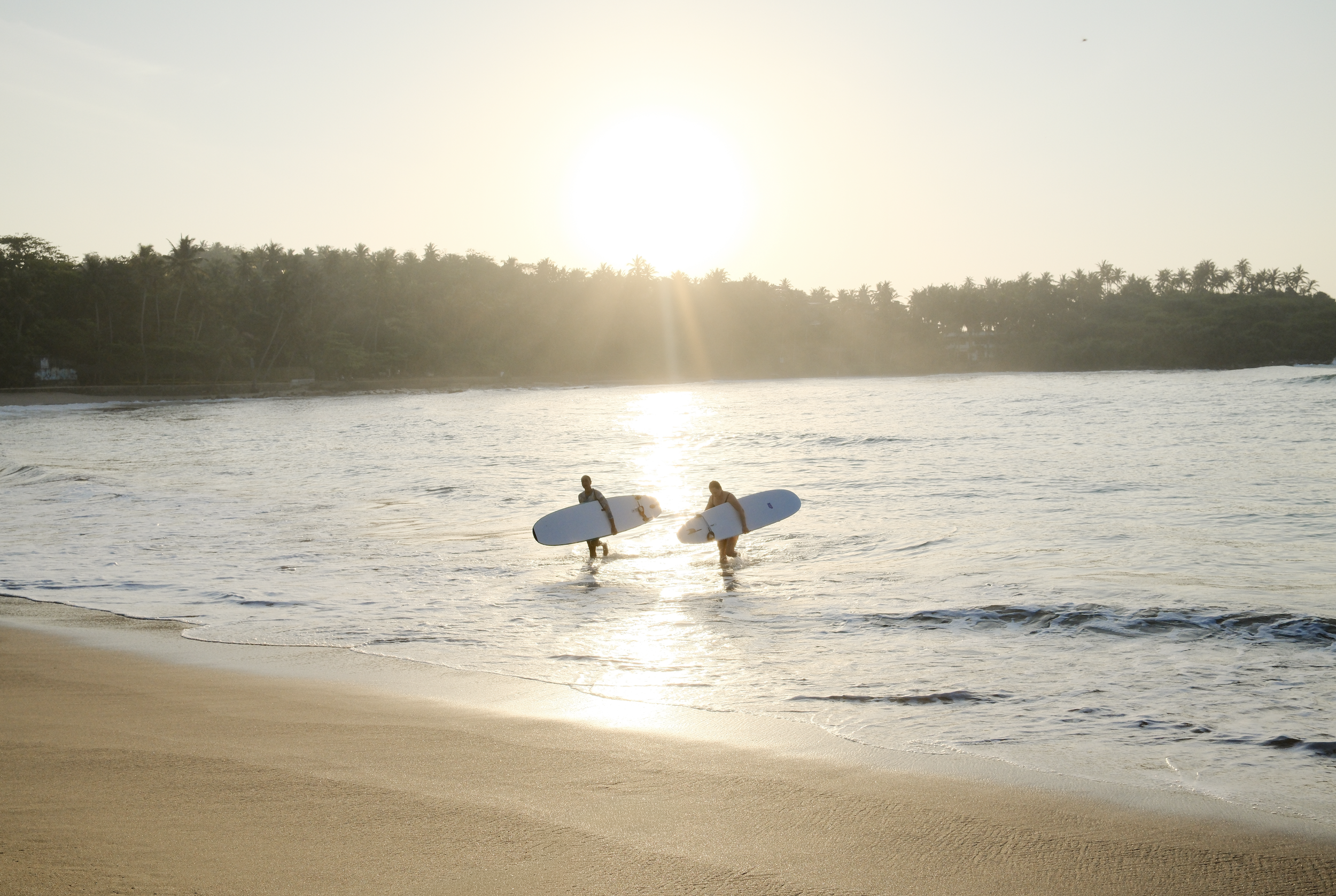 Two surfers carrying surfboards walking into the water at a beach during sunset or sunrise.