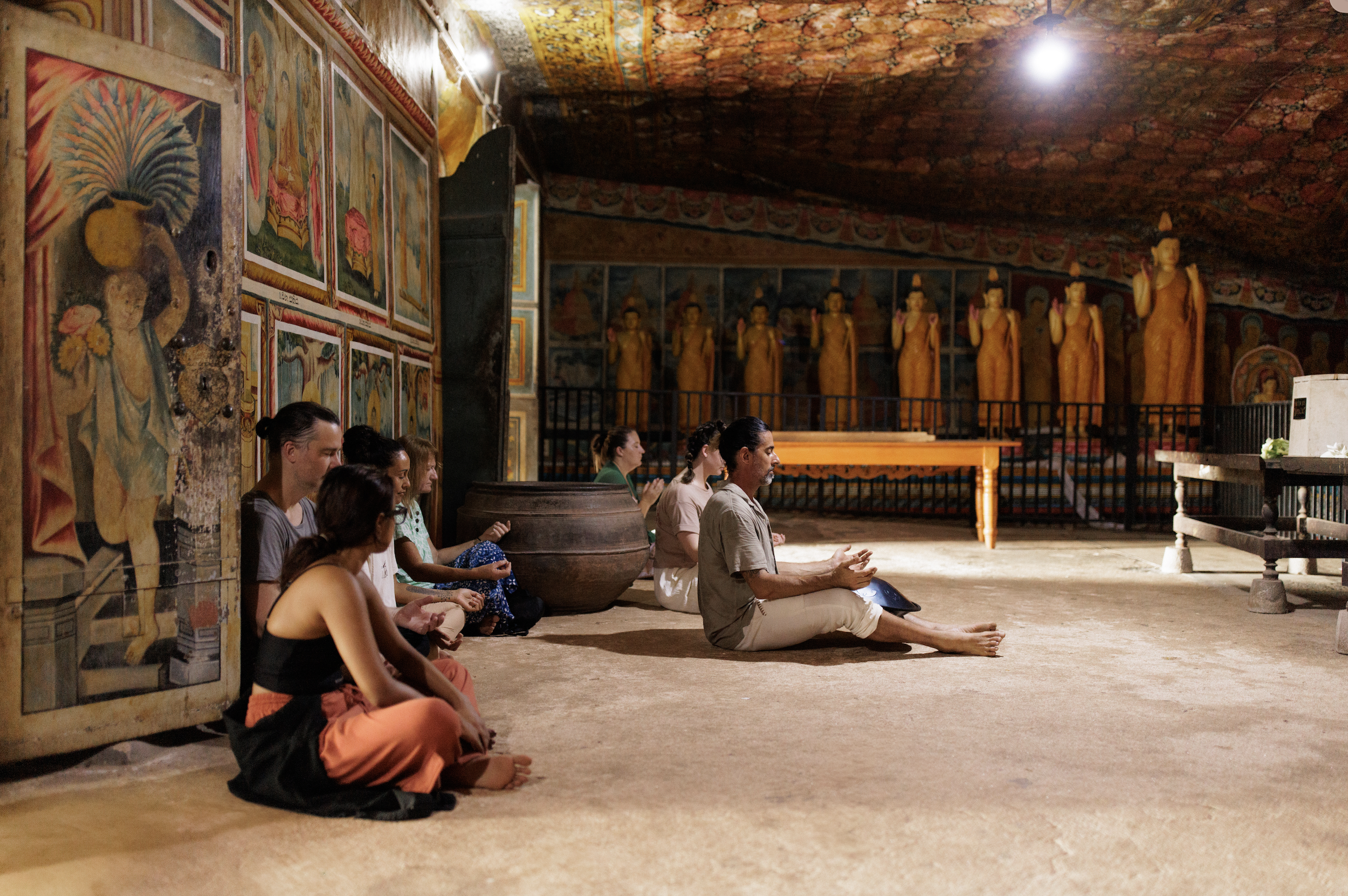 A group of people sitting cross-legged and meditating inside a temple with colorful artwork and statues of standing Buddhas on a wall in the background.