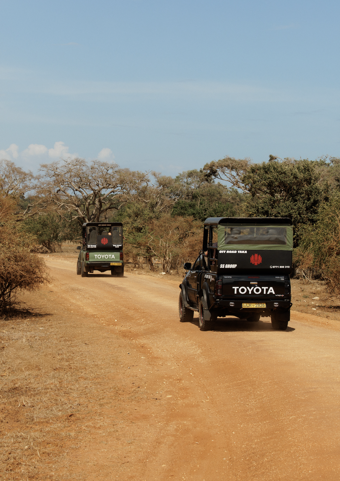 Two black Toyota off-road vehicles driving on a dirt road through a dry, bushy landscape with sparse trees under a blue sky.