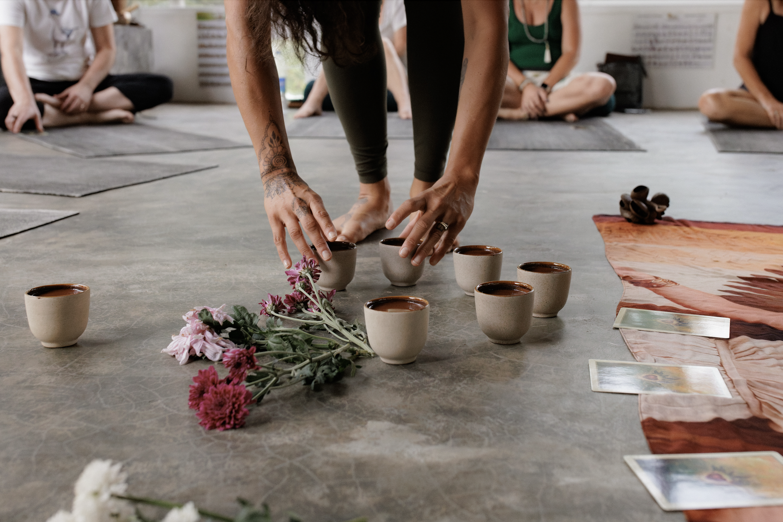 A person setting up a ceremony with beige cups, flowers, and tarot cards on a floor, while others sit on mats in the background.
