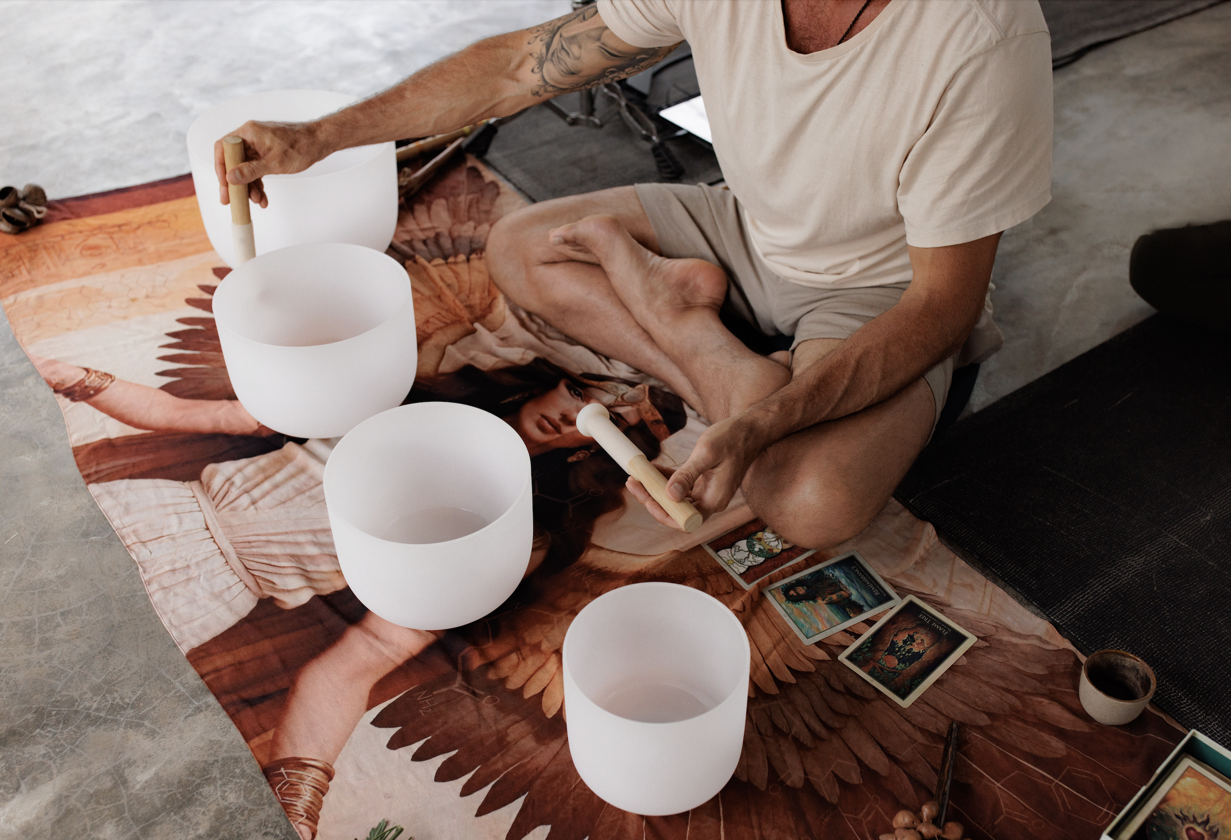 Person sitting cross-legged on a bed, playing crystal singing bowls with tarot cards and a small cup nearby during a spiritual or musical session.