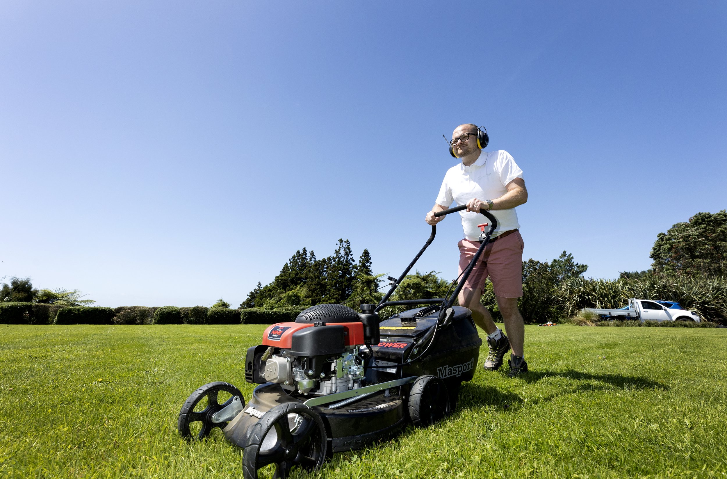 A man wearing a white shirt, pink shorts, and headphones is using a lawn mower on a grassy field under a clear blue sky.