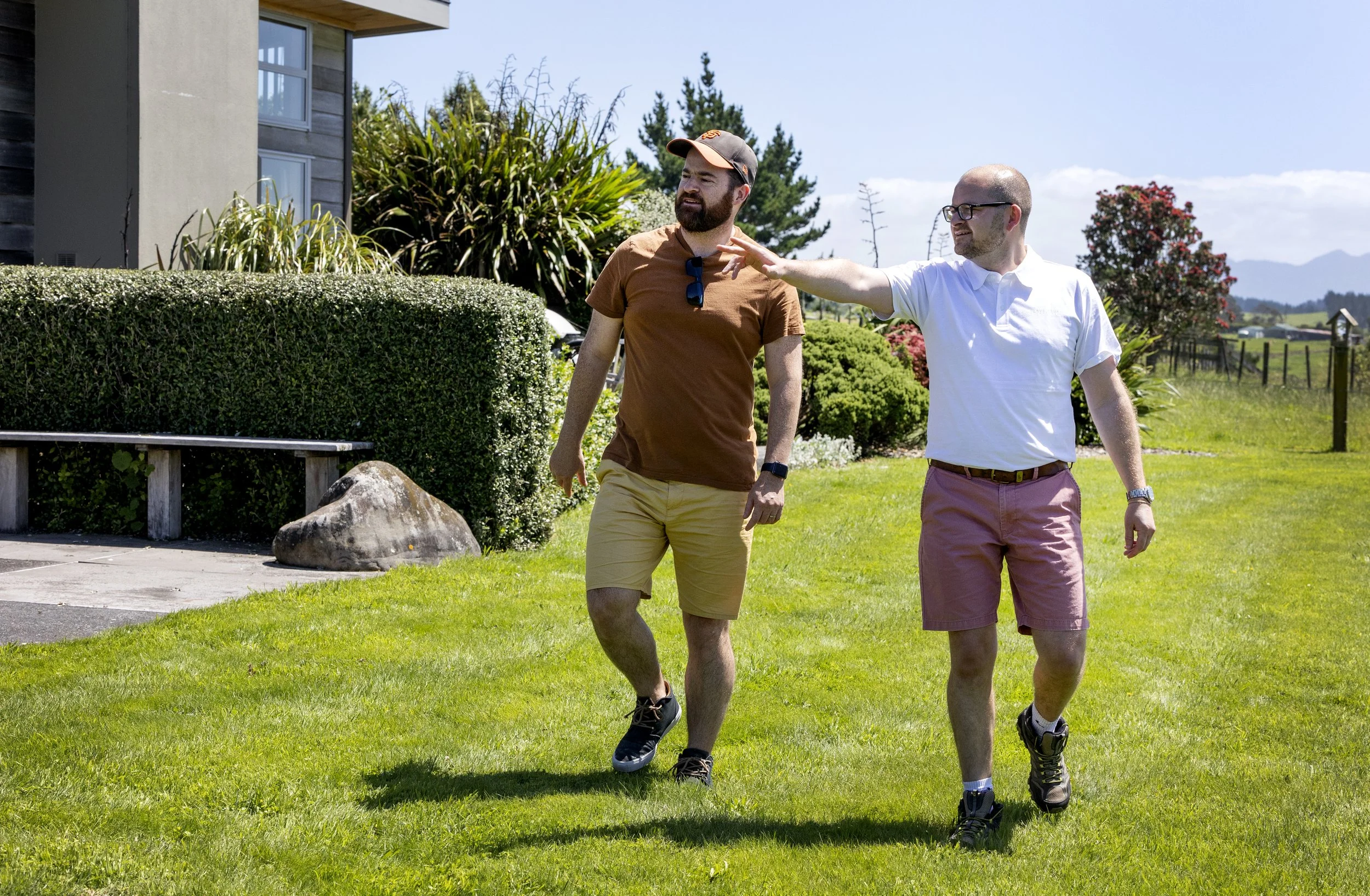 Two men walking on a grassy lawn, one is reaching out to the other while talking, with a house and trees in the background.
