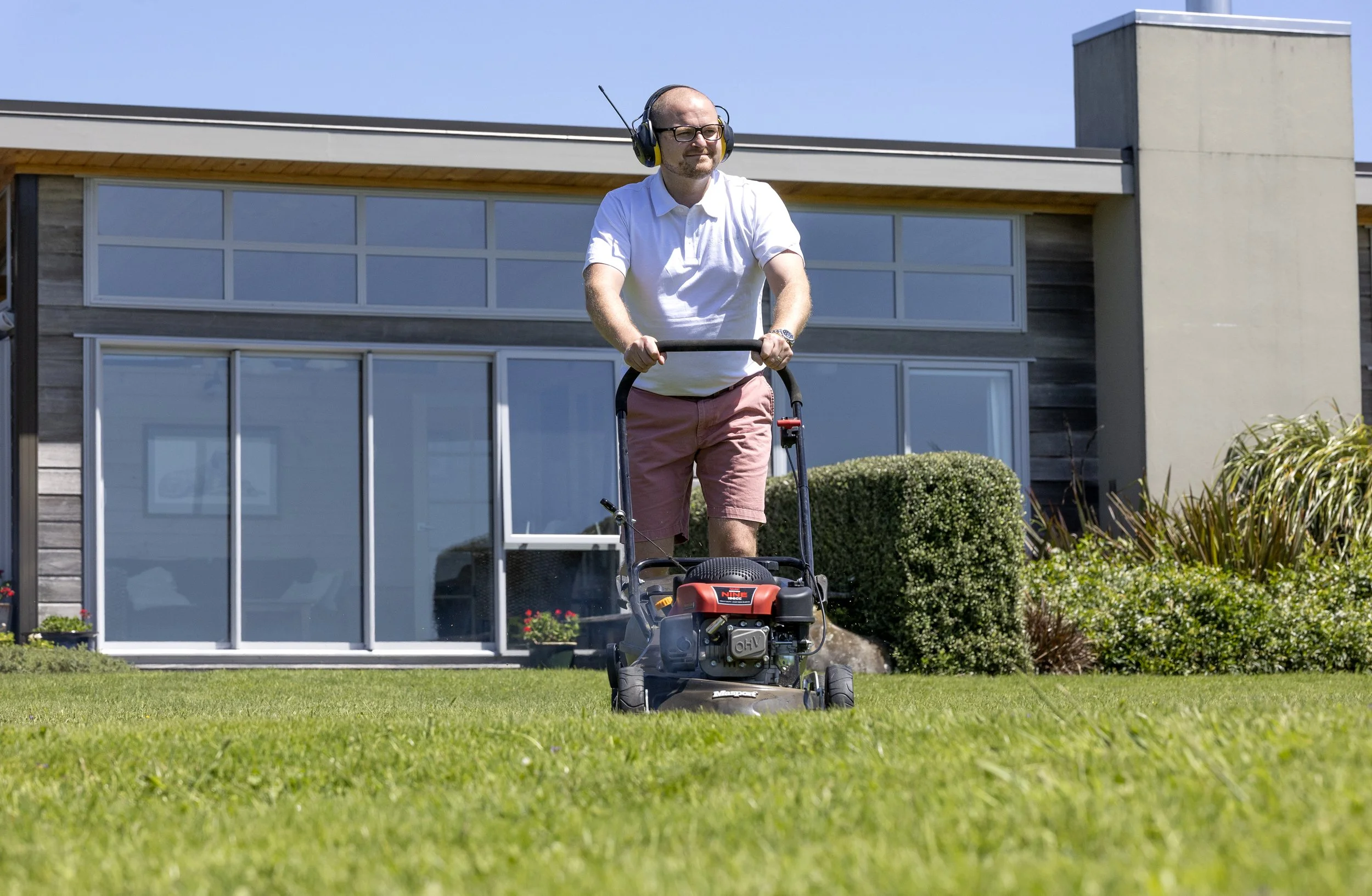 A man wearing a white shirt and pink shorts mowing the lawn with a gasoline-powered lawnmower in a backyard of a modern house with large windows and well-maintained bushes.